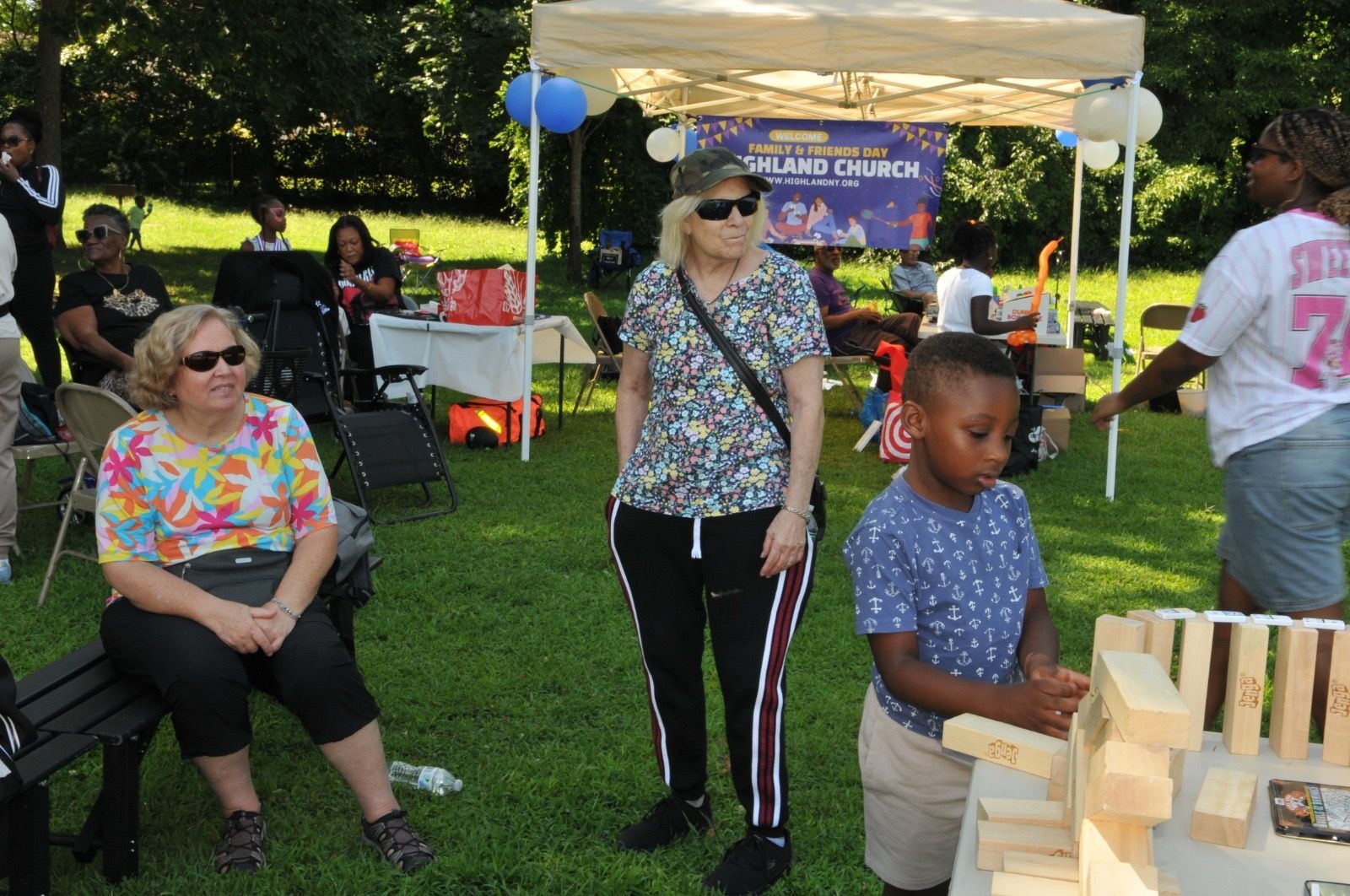 People seated and standing at an outdoor park event with a child playing at a wooden game table.