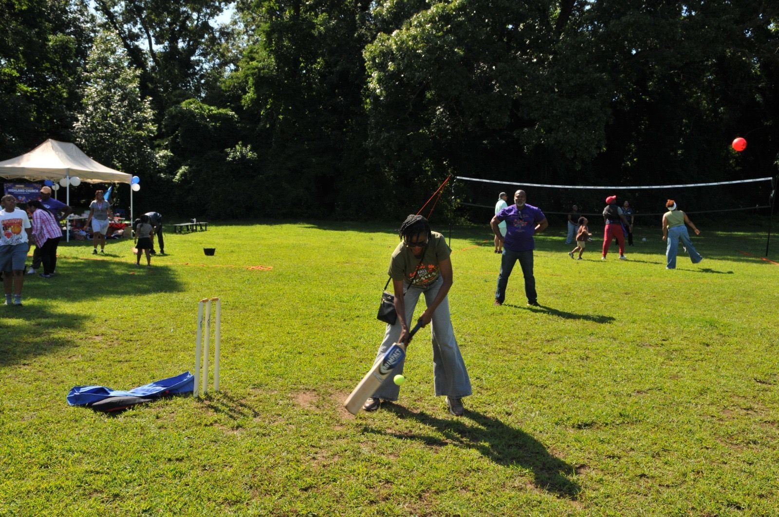 People playing cricket on a sunny grassy field with trees and a tent in the background