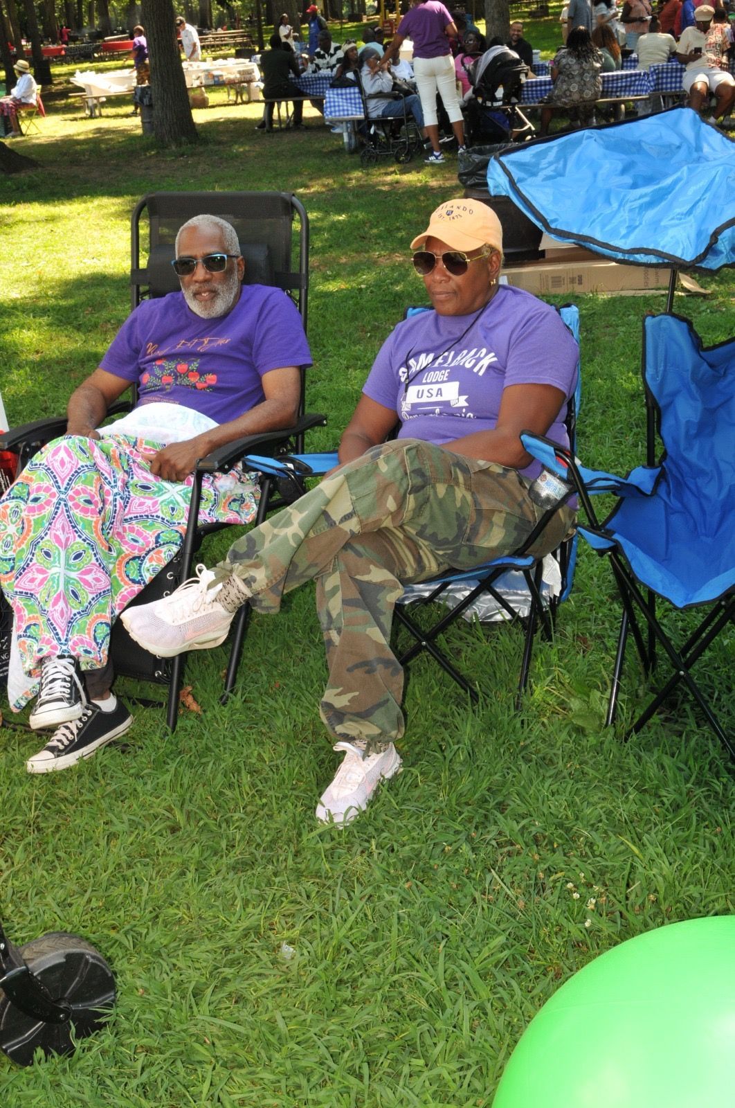Two people in matching purple shirts sit in folding chairs on a grassy park lawn during an outdoor gathering