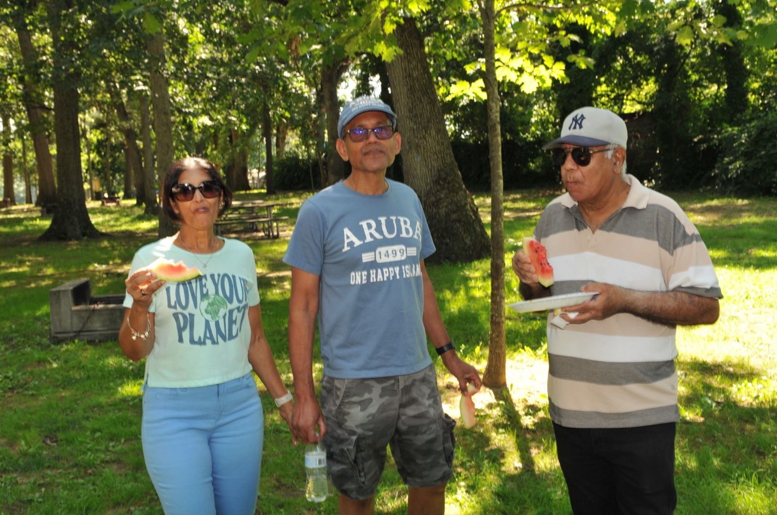 Three people standing outdoors in a wooded park, holding cups and wearing casual clothes.