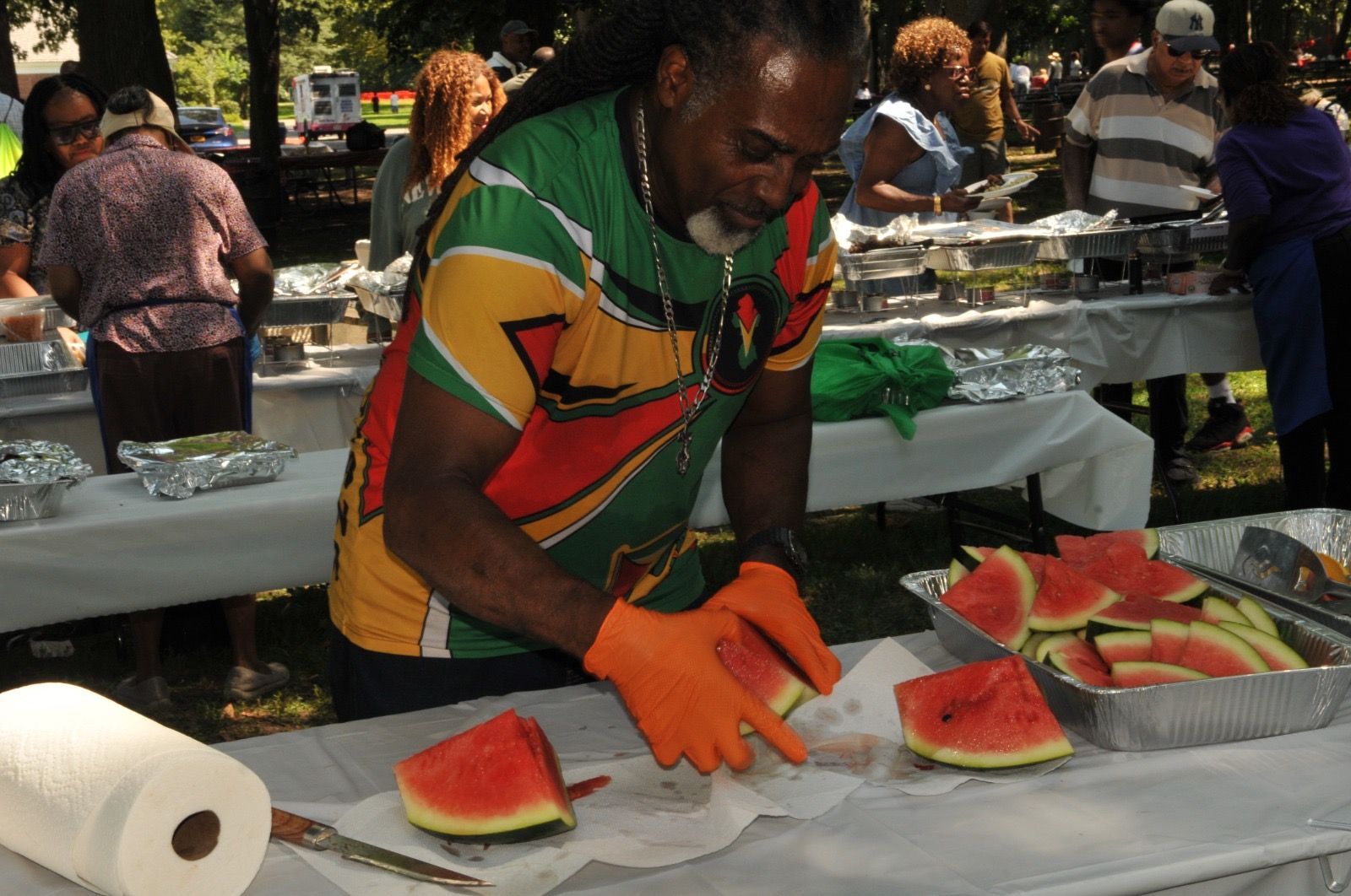 Man slicing watermelon at an outdoor food stall with orange gloves and a colorful shirt