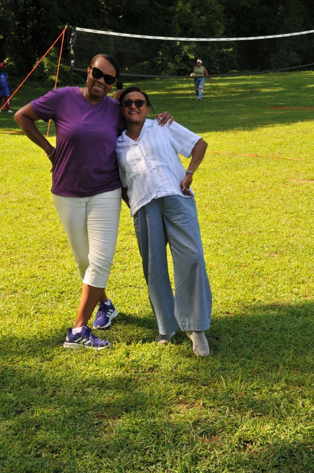 Two people posing together on a sunny grassy field, one in a purple shirt and white pants, the other in a white shirt and blue jeans.