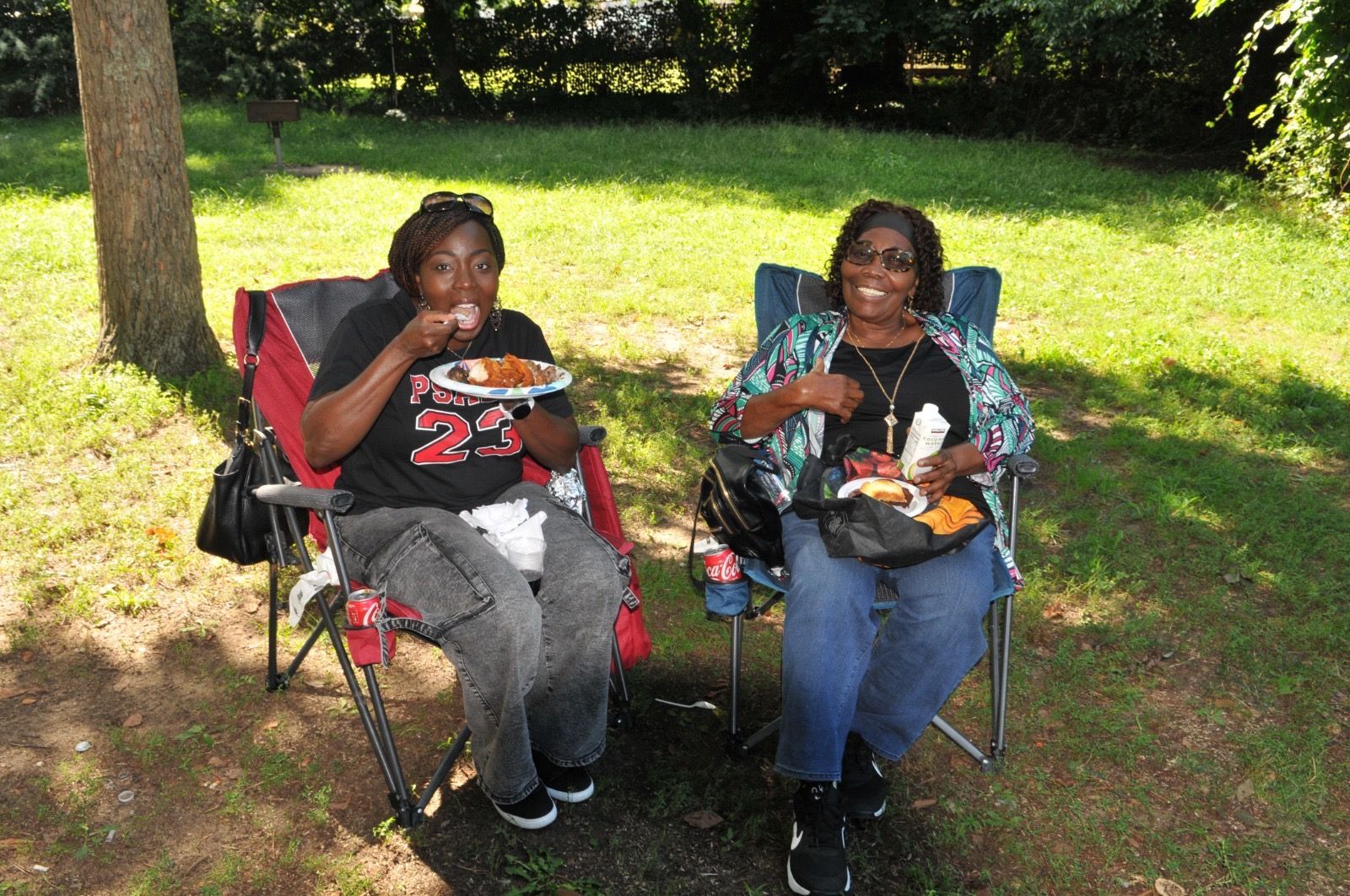 Two people sitting in folding chairs on a sunny lawn, eating food and smiling at the camera.