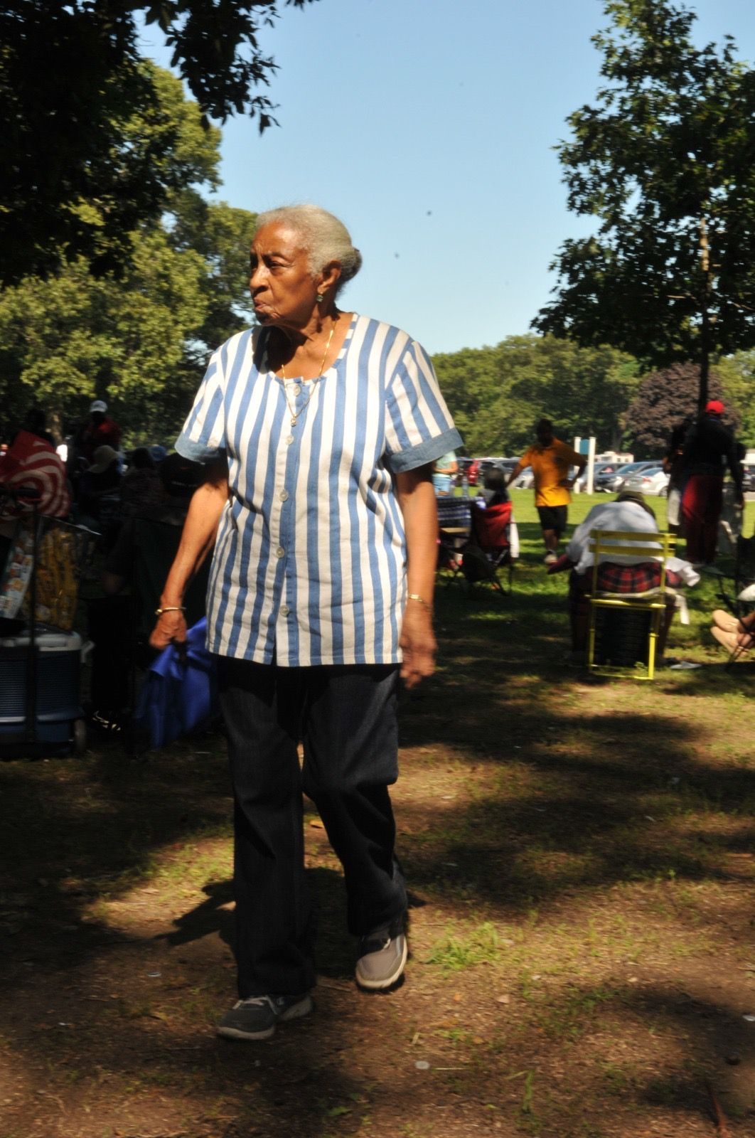 Person in a striped shirt walking in a sunny park with people and trees in the background