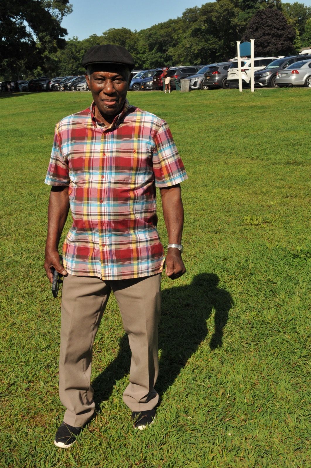 Man standing on a grassy field in a plaid shirt and hat, with parked cars and trees in the background