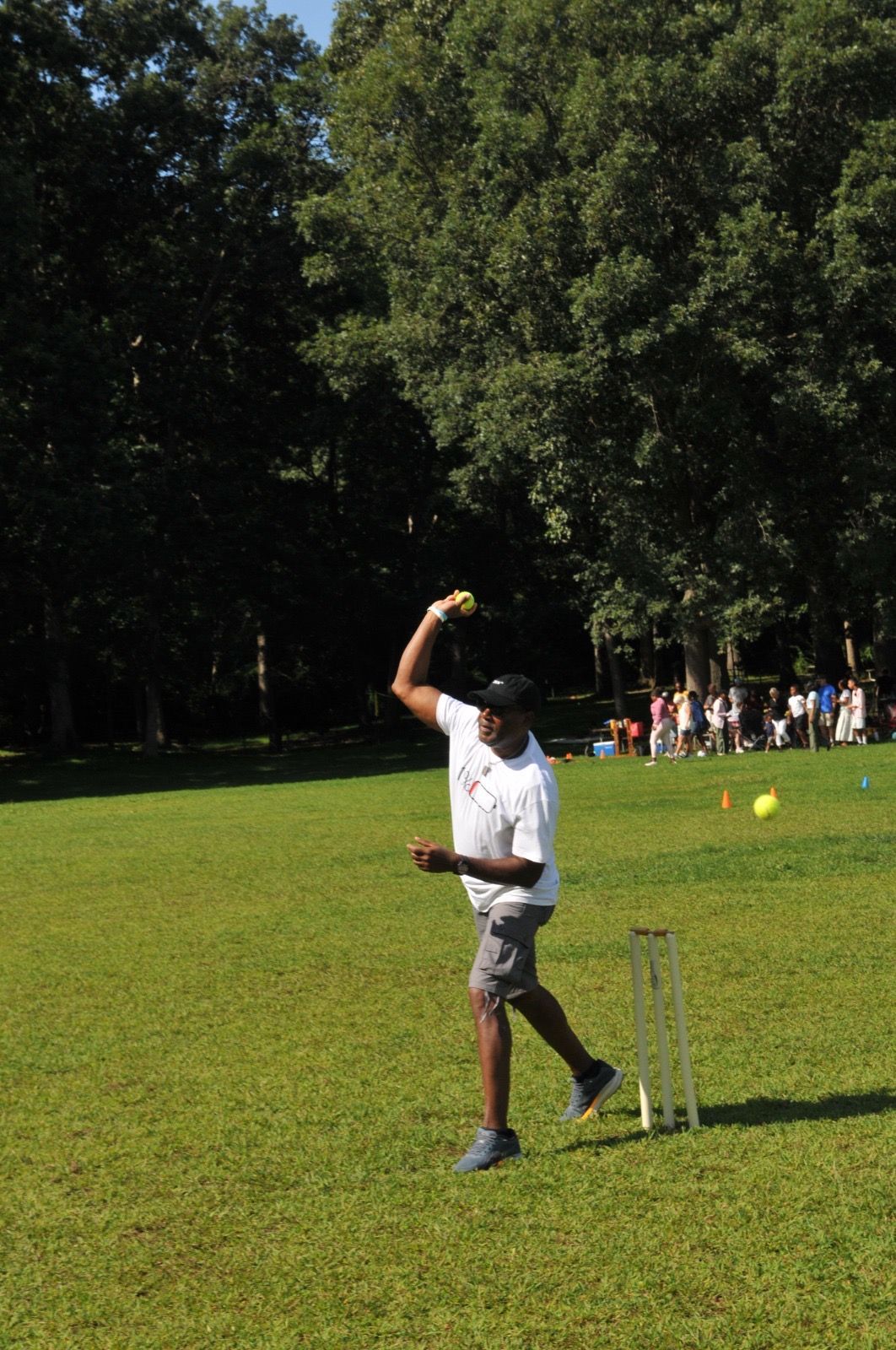 Cricketer in white uniform bowling on a grassy field with a wicket nearby, trees and spectators in the background