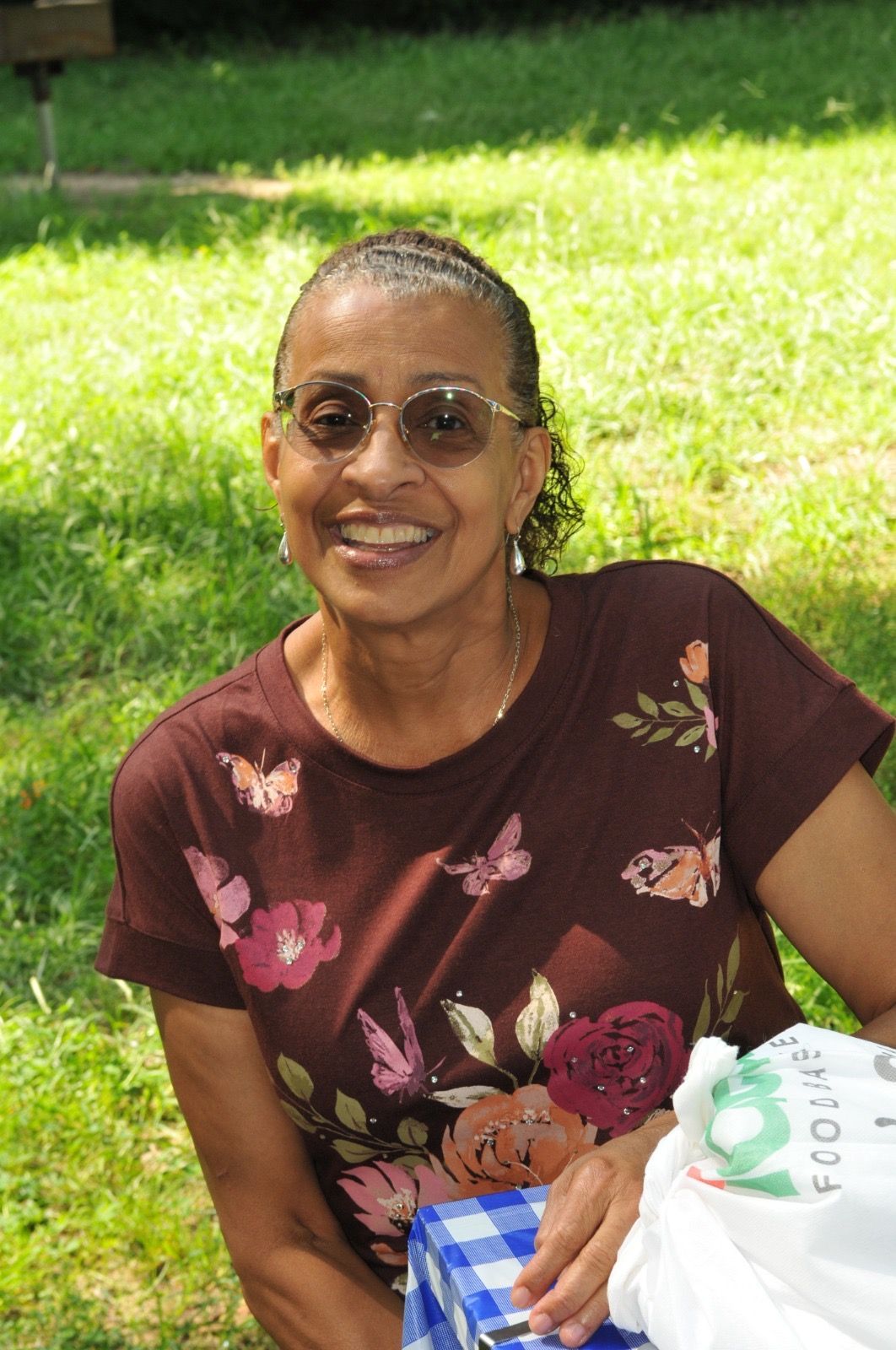 Smiling person seated on grass outdoors, wearing sunglasses and a brown floral shirt.