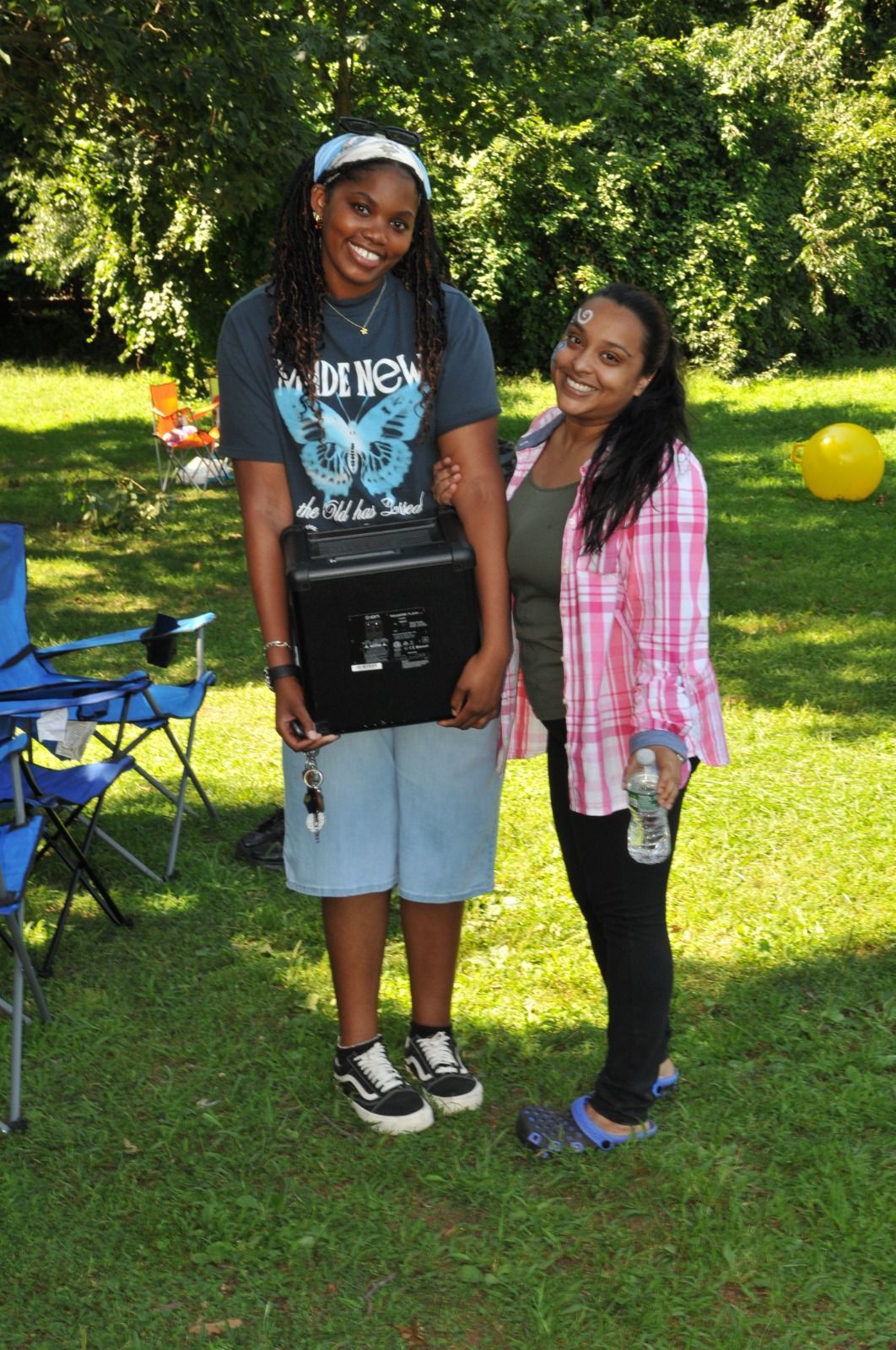 Two smiling people standing on grass in a sunny park, one holding a water bottle.