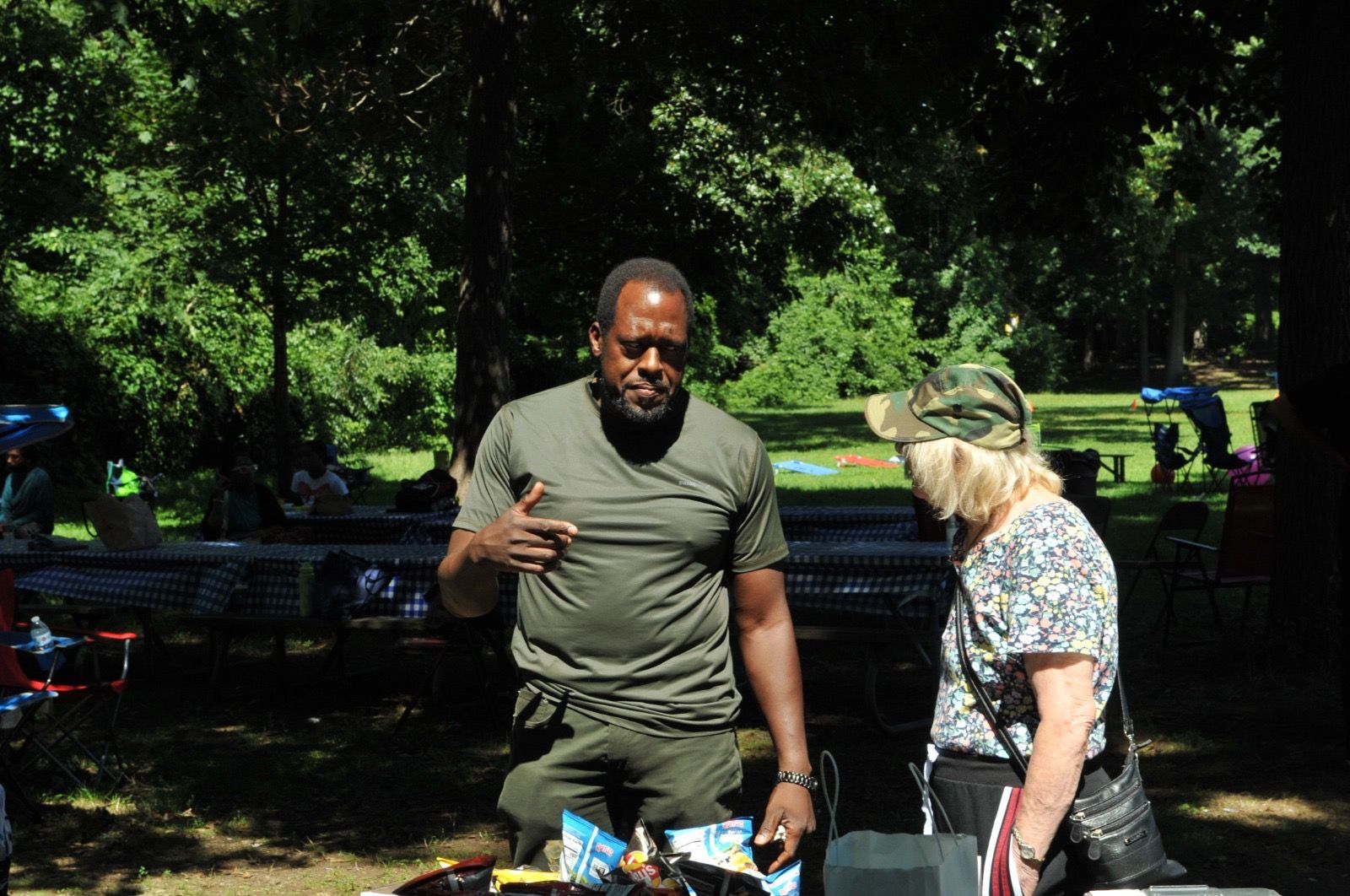 Two people talking at a picnic table in a sunny park, with trees and benches in the background.