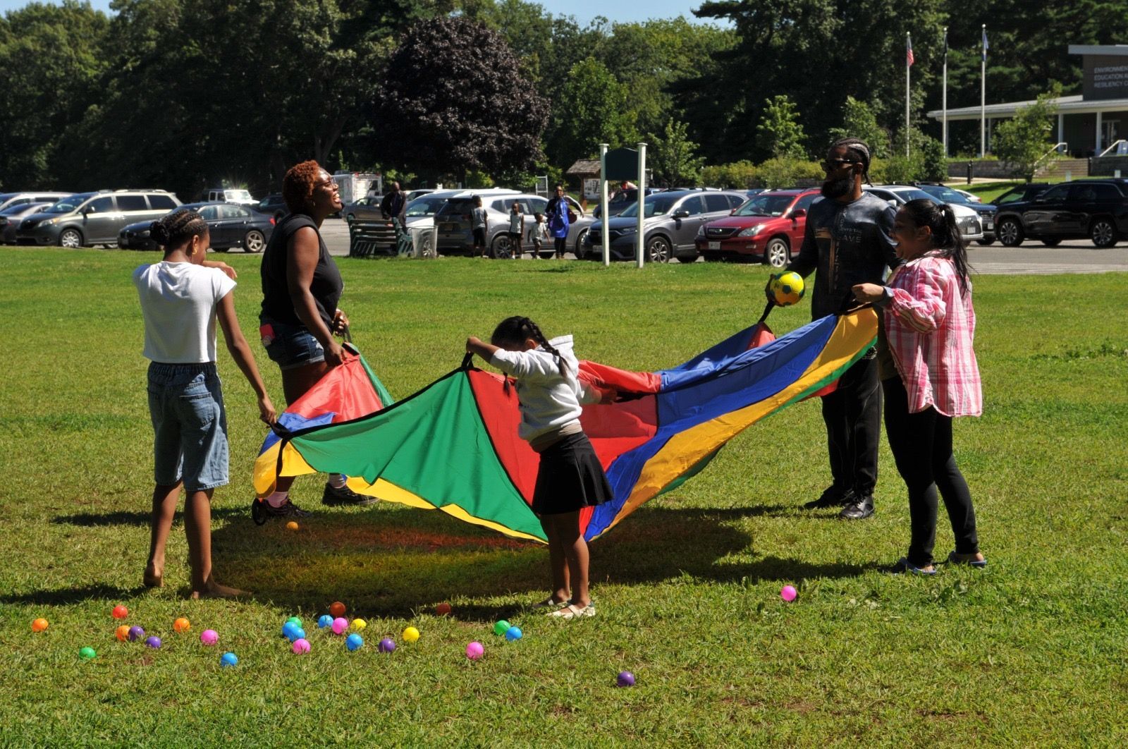 Children and adults hold a colorful parachute on a grassy field with balloons scattered nearby