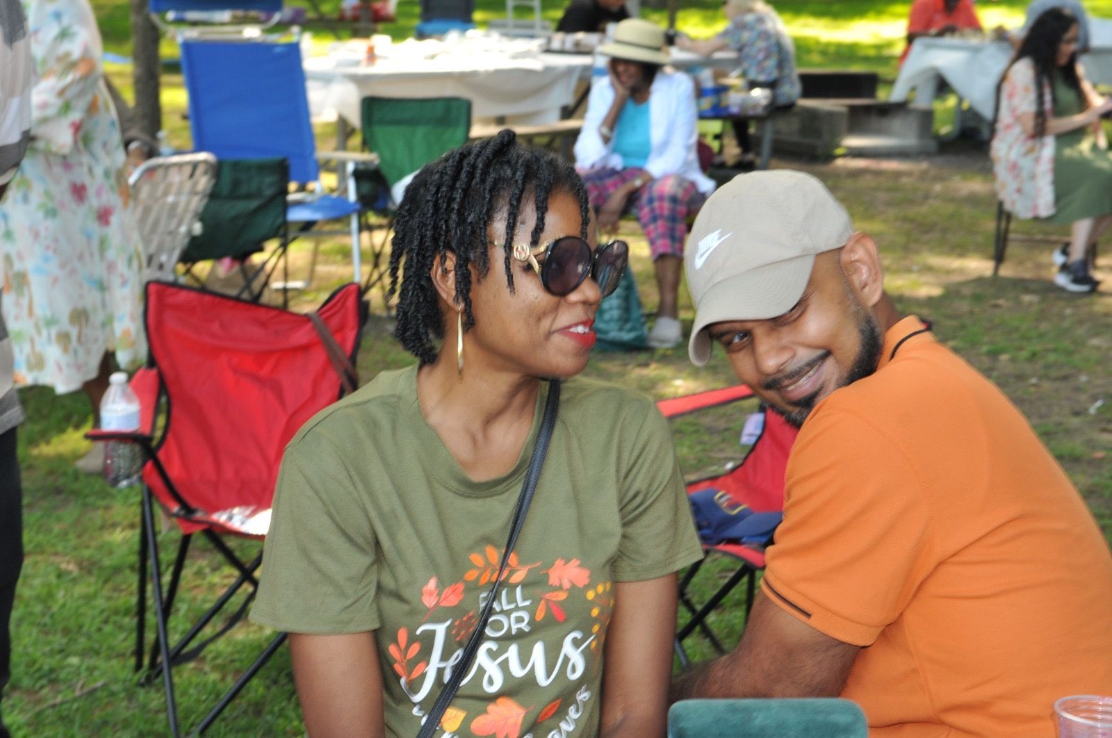 Two people sit close together at an outdoor gathering, smiling in a park with folding chairs and trees behind them.