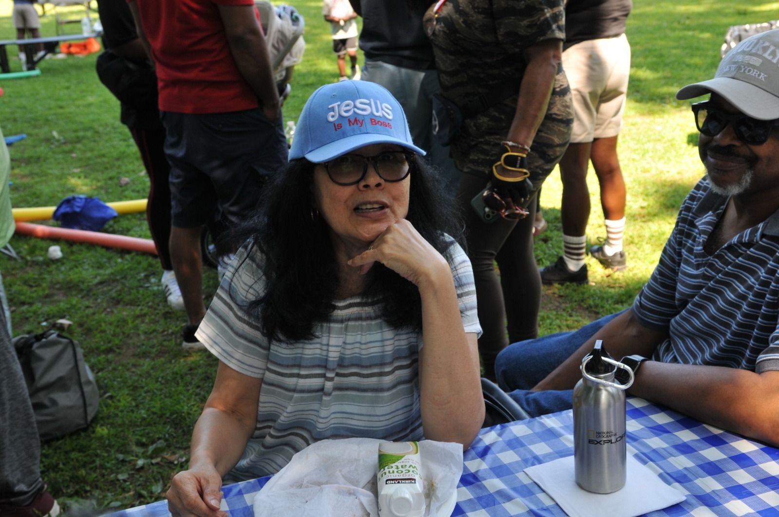 Woman in blue cap chatting at a picnic table outdoors, with other people standing nearby.