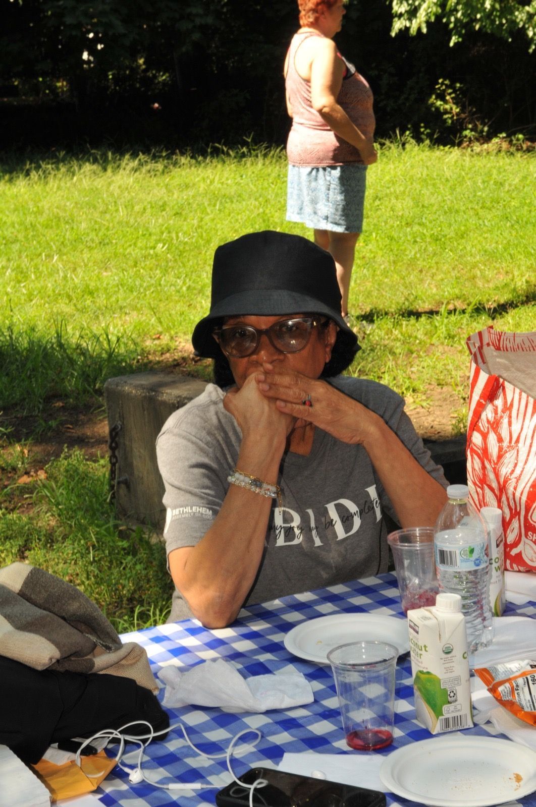Person in a black hat sits at a picnic table with a blue checkered tablecloth outdoors.