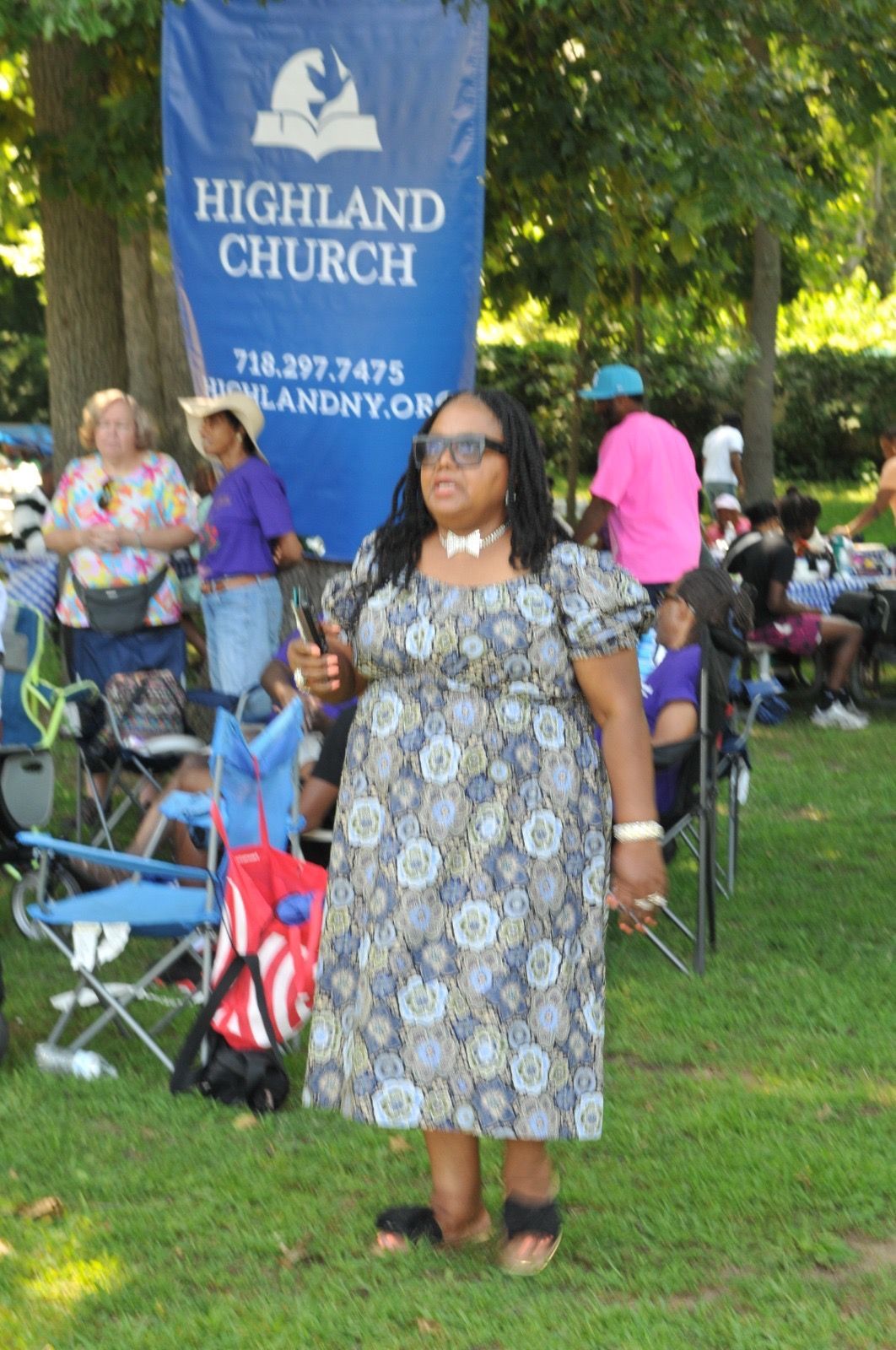Woman standing on grass at a Highland Church outdoor gathering, with people and a blue church sign behind her.