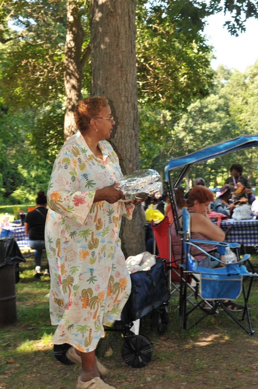Woman in a floral dress standing in a park beside seated people under a blue canopy.