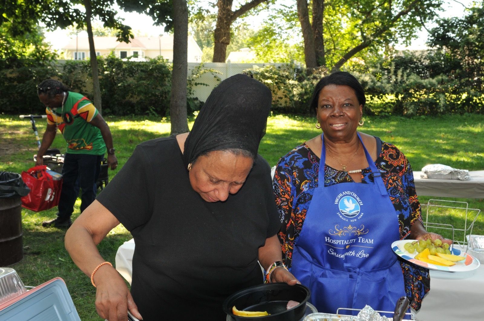 Two women serving food outdoors at a community gathering, one leaning over a dish, the other holding a plate.