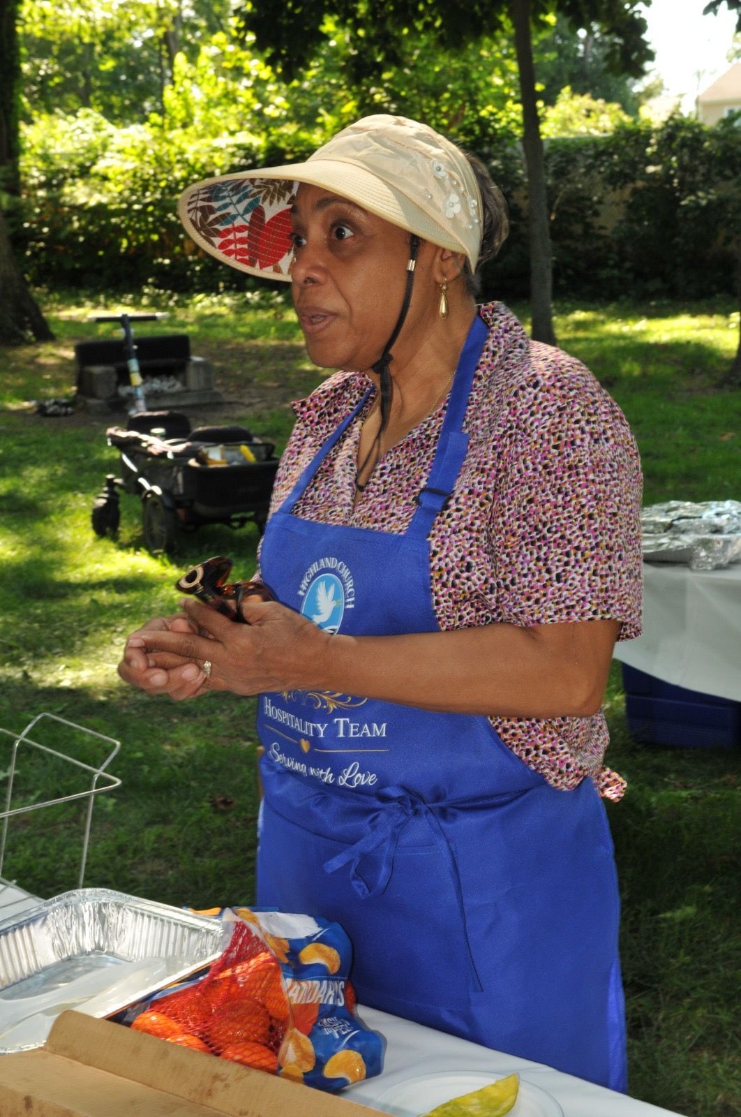 Woman in blue apron and sun hat preparing food at an outdoor picnic table.