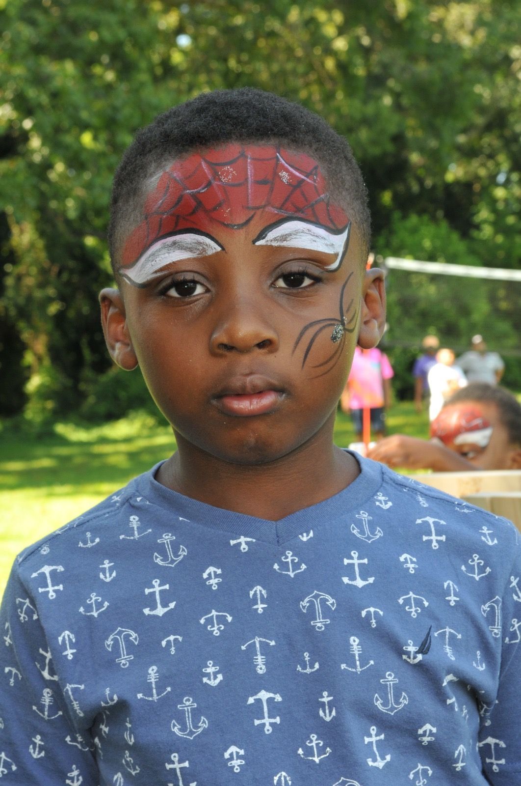 Child with face paint and a blue patterned shirt standing outdoors, looking at the camera