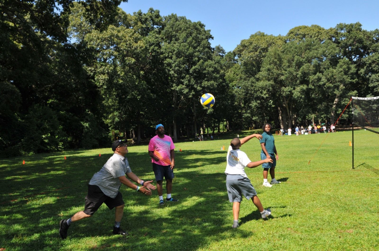 People playing volleyball on a sunny grassy field with trees in the background