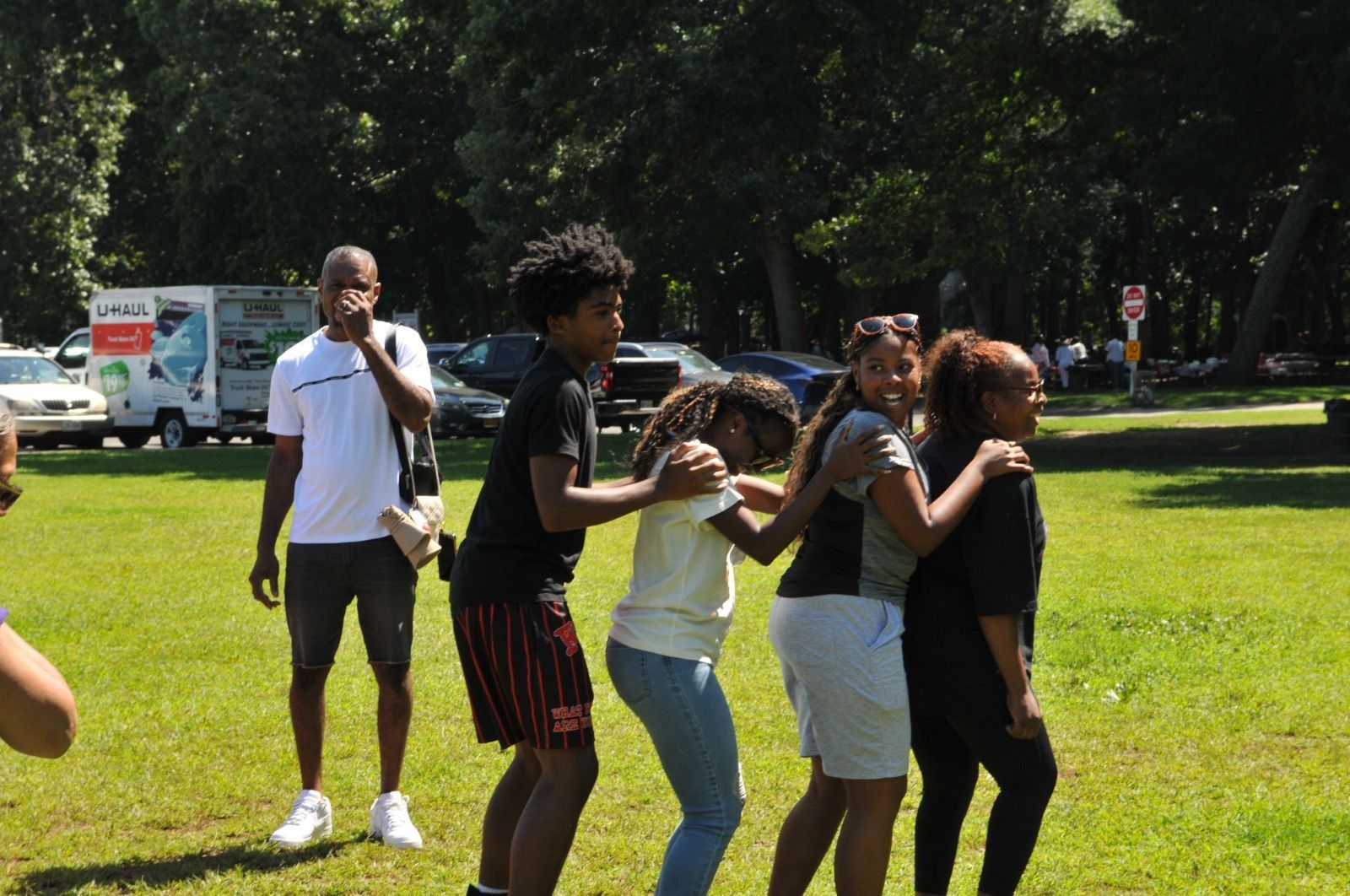 People playing a game on a grassy park field, with one person standing nearby and a van in the background