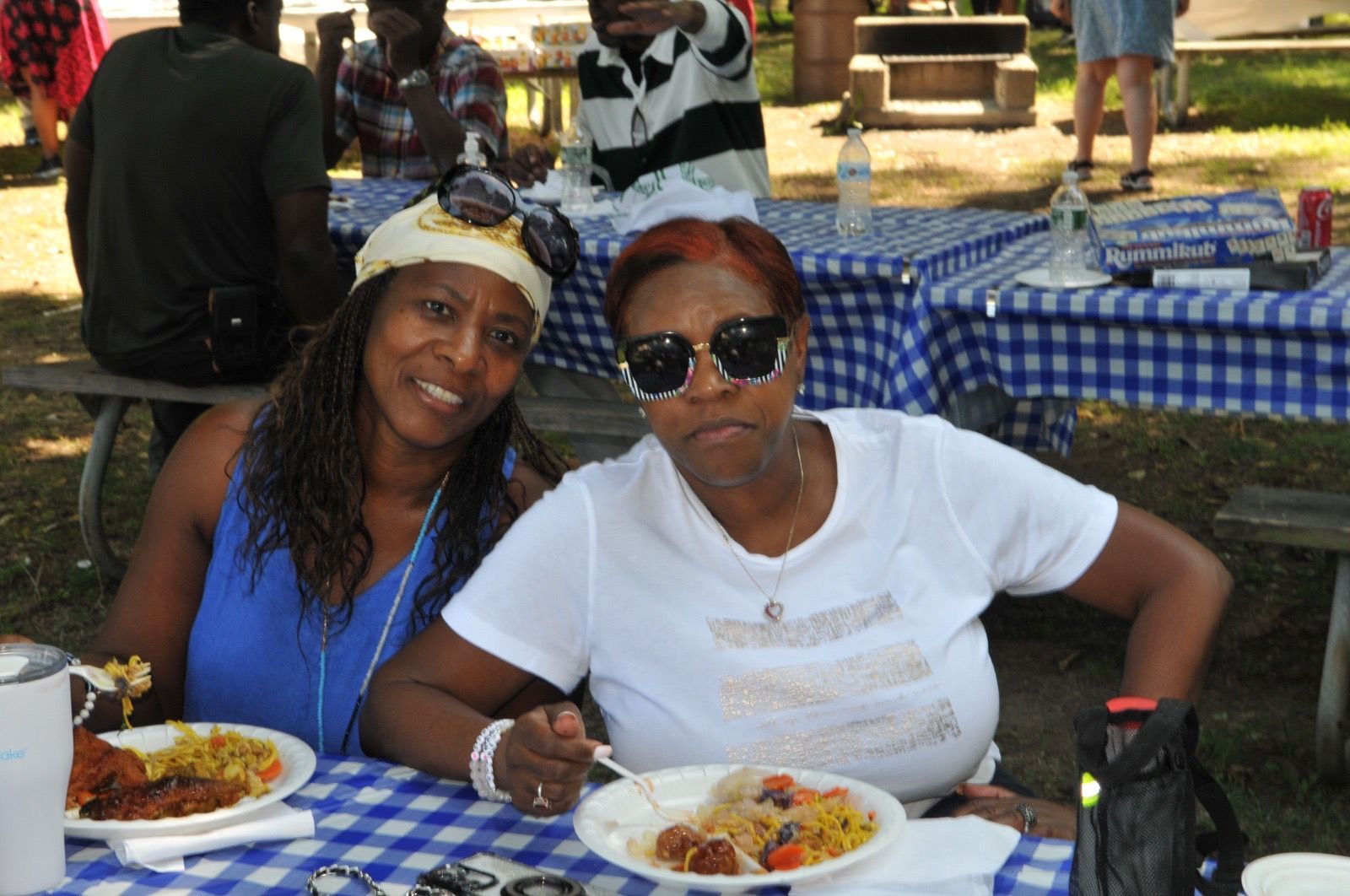 Two people seated at a picnic table with plates of food under a blue checkered canopy