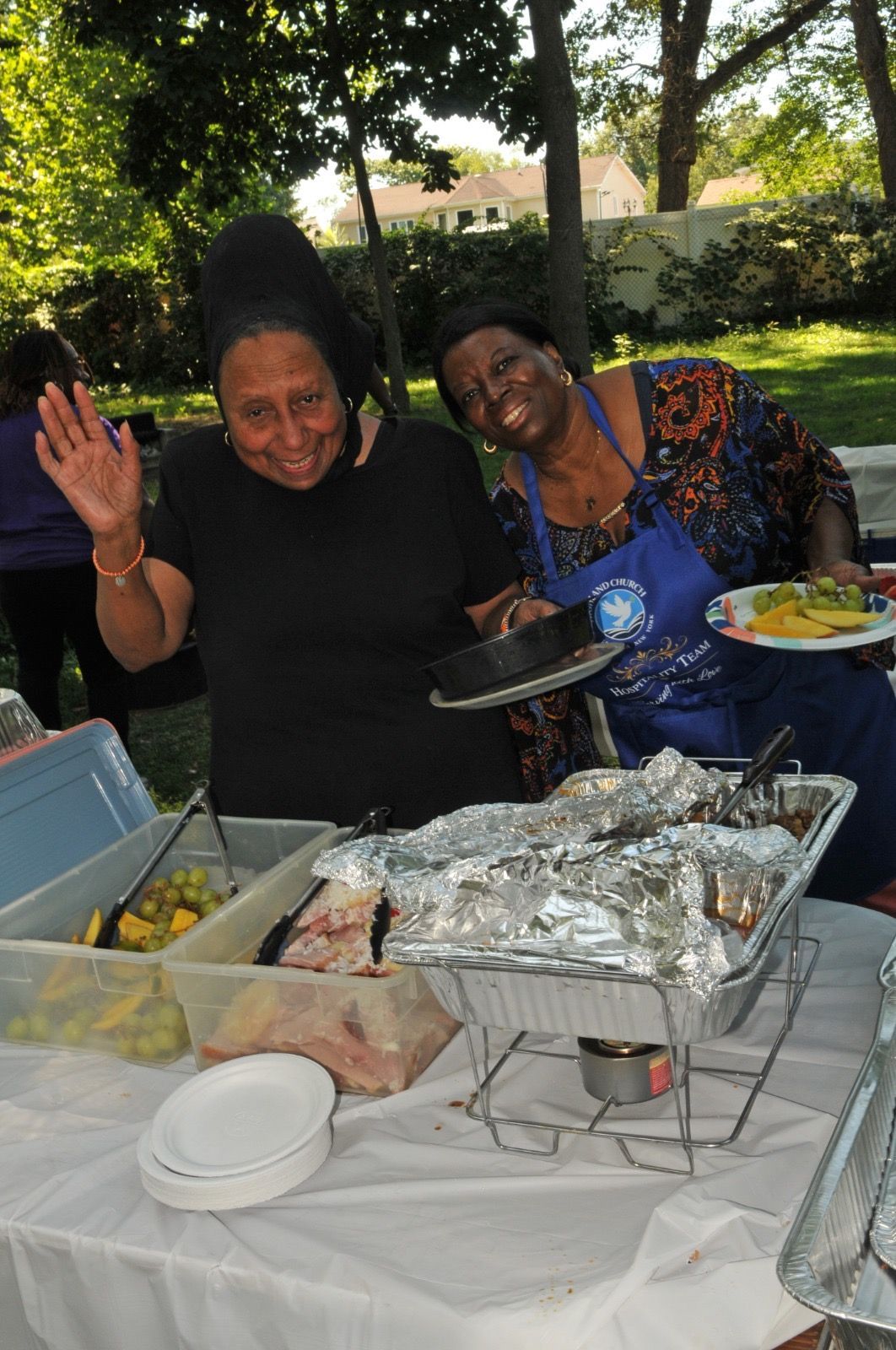 Two people smiling and waving at a picnic table with trays of food outdoors.