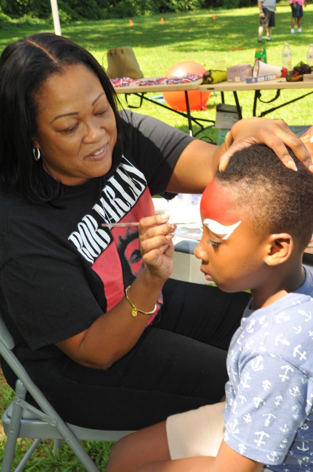 Woman painting a child’s face at an outdoor event, seated in folding chairs.