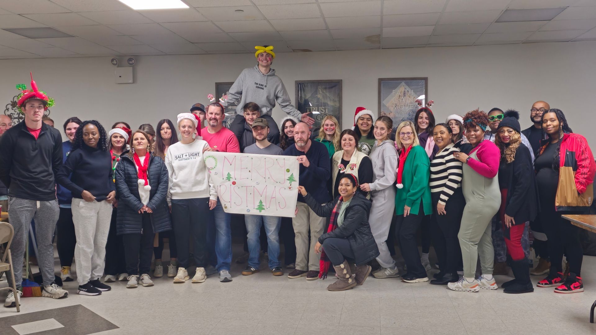 Large group posing indoors, holding a decorated poster during a festive holiday gathering. Christmas Toy Drive 2025.