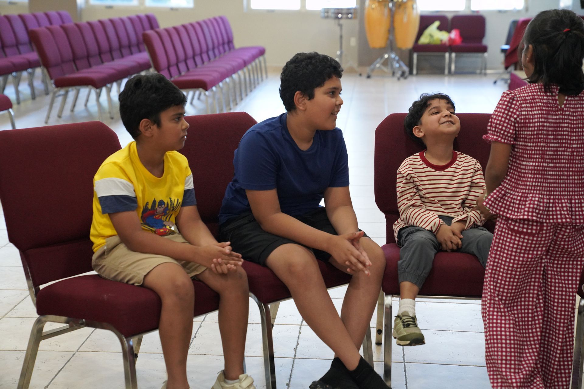 Children sitting and chatting in a bright room with rows of red chairs behind them