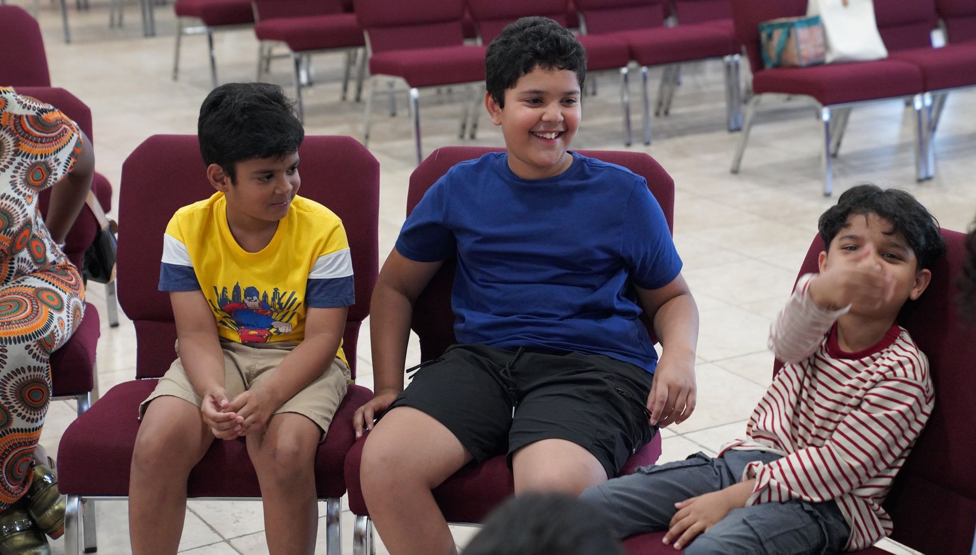 Three children sitting on chairs in a hall, smiling and chatting.