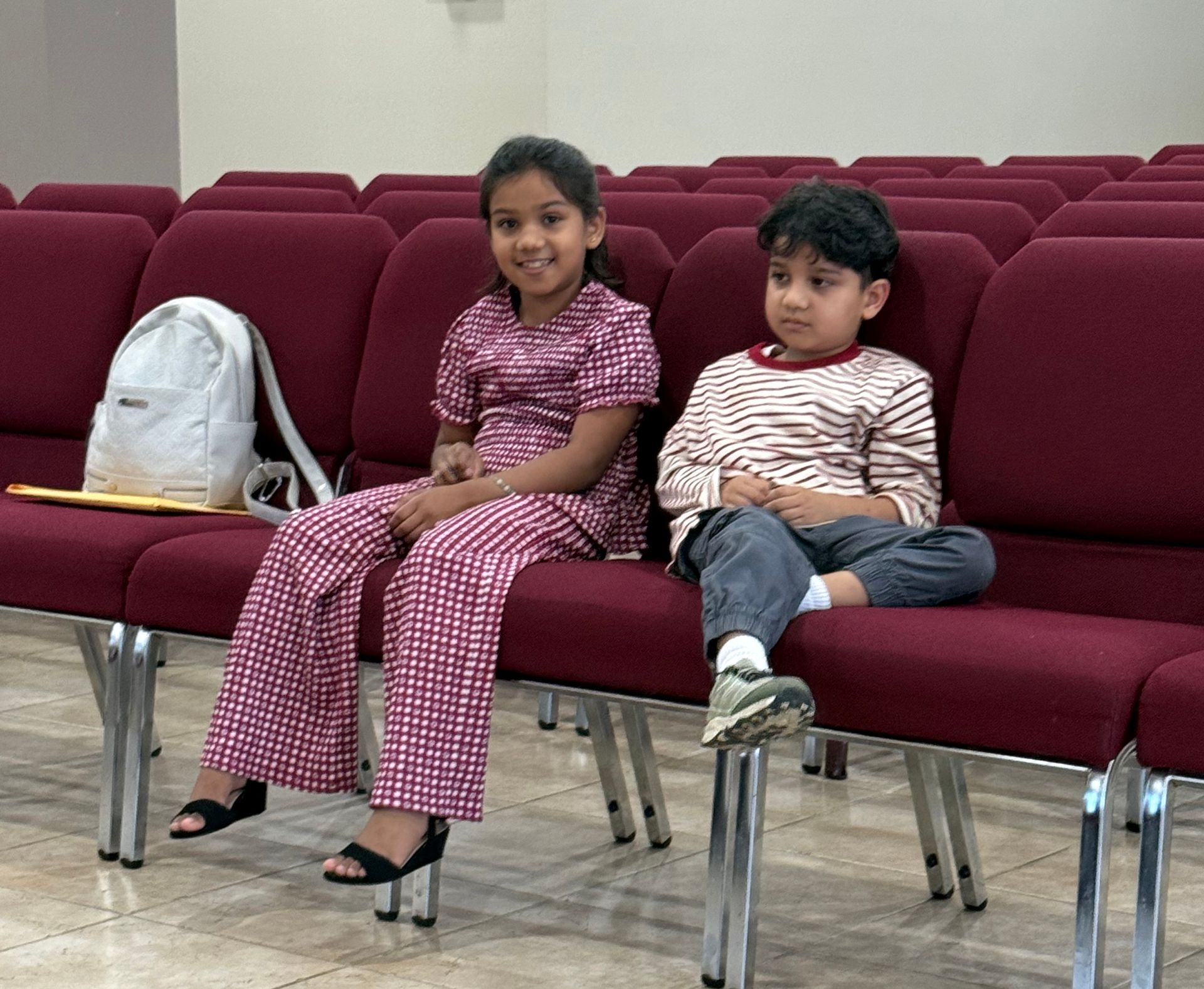 Two children sitting on maroon auditorium chairs, one smiling, with a white backpack beside them.