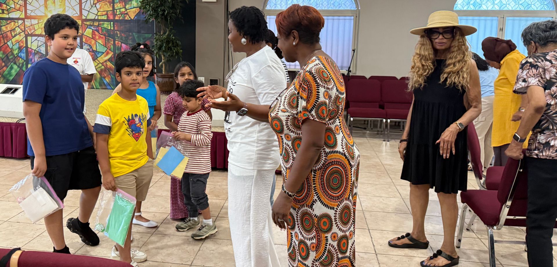 Children and adults standing in a room, holding papers and chatting during an indoor gathering