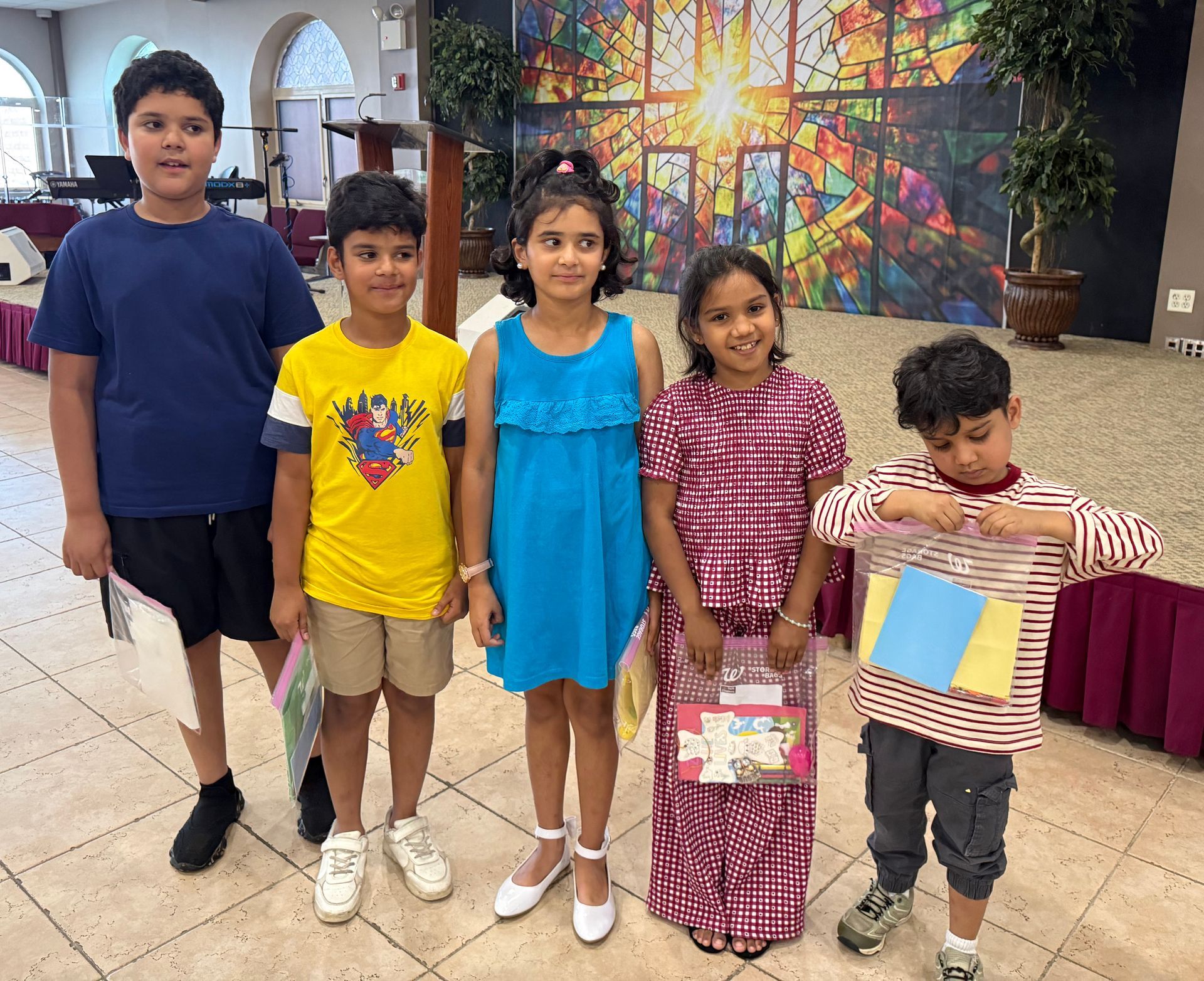 Five children posing indoors, holding colorful papers in a bright lobby with a stained-glass mural.