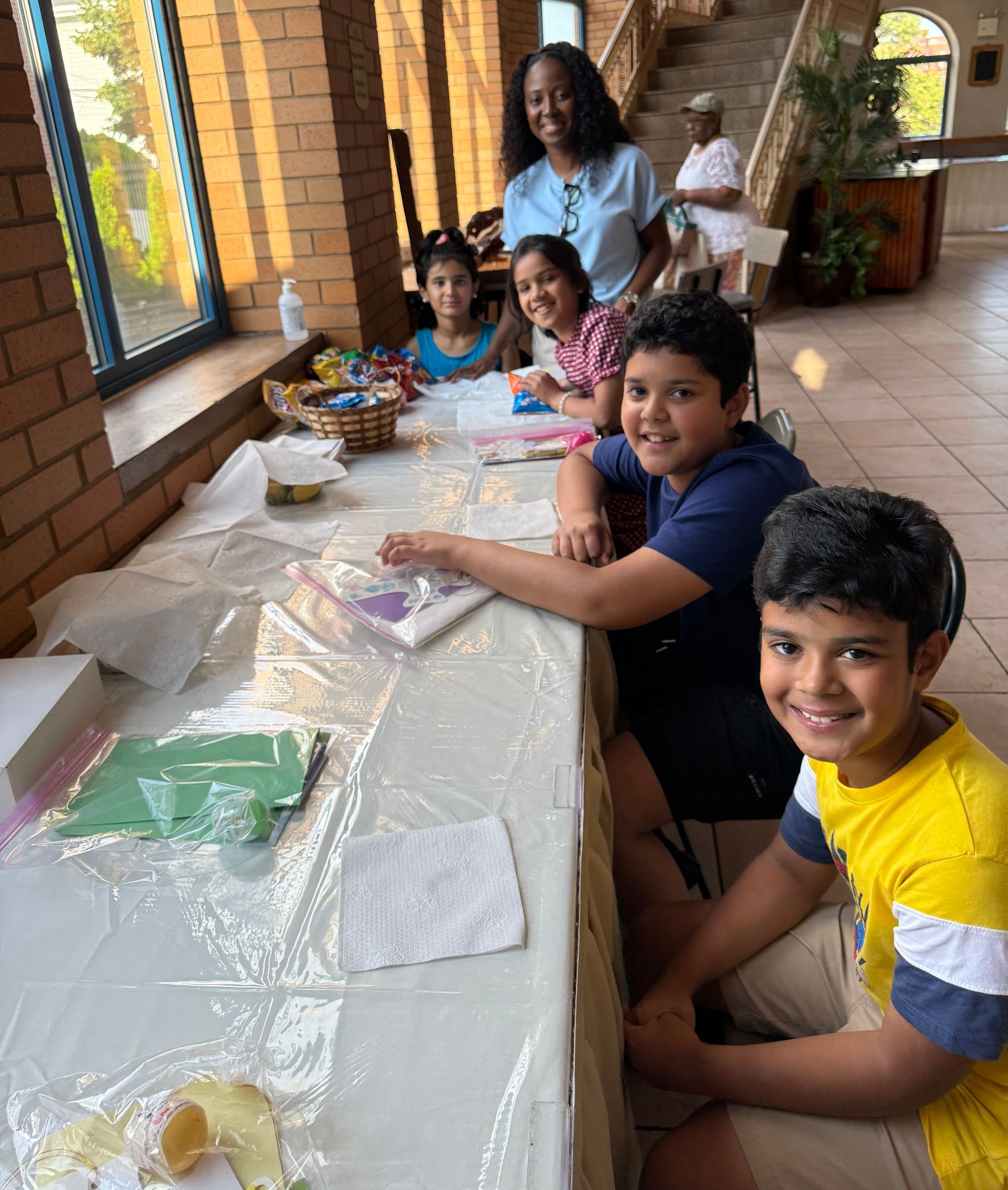 Children seated at a long table with craft supplies in a bright indoor hallway