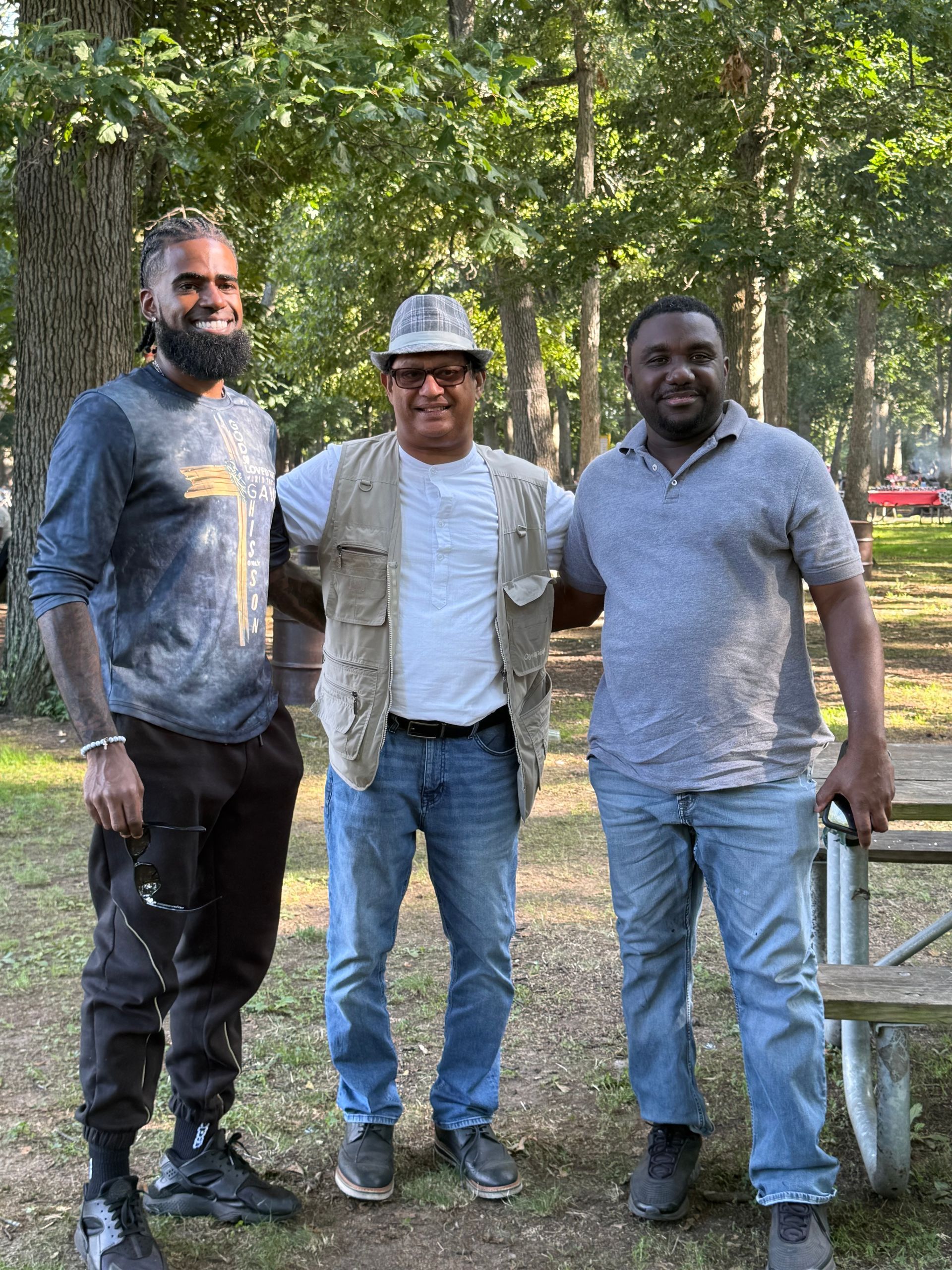 Three men standing together outdoors in a wooded area, smiling at the camera.