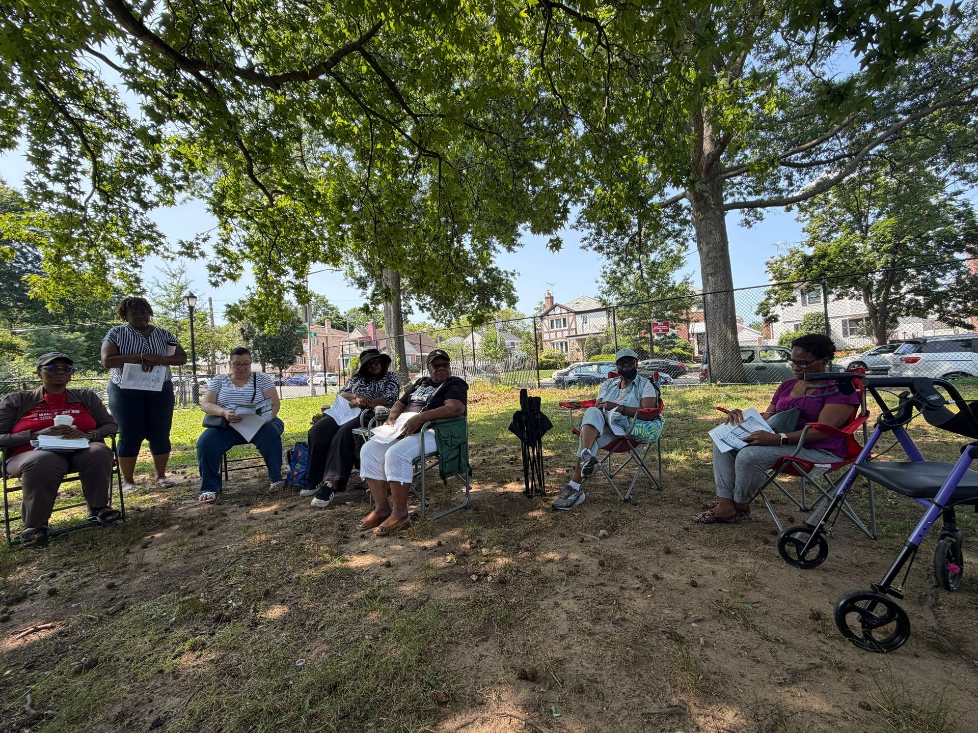 People seated outdoors in a shaded park under large trees, with bicycles nearby.