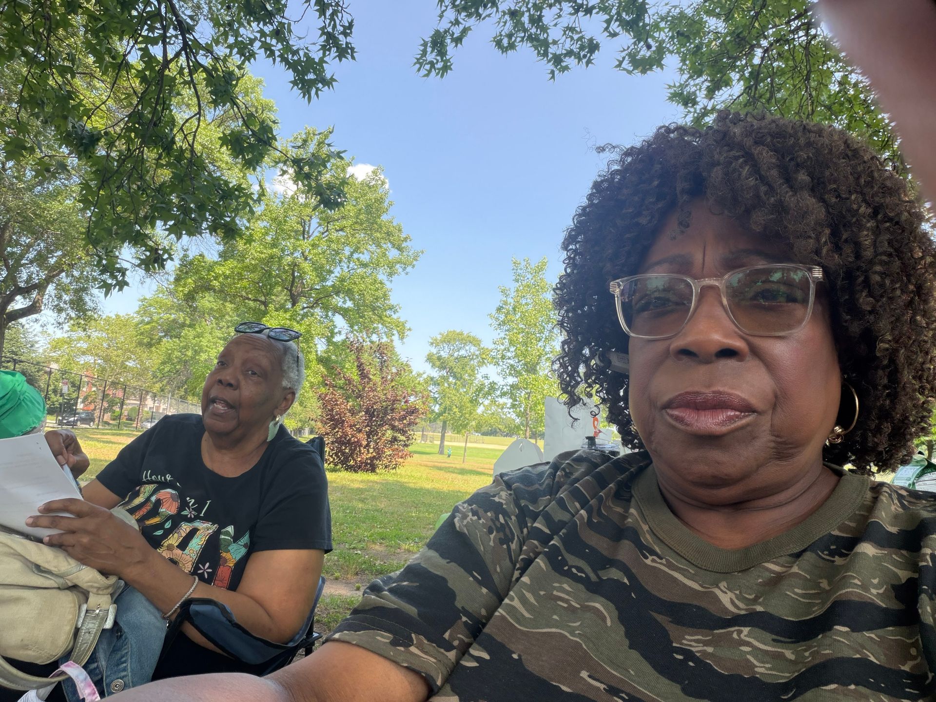 Two people sitting outdoors under trees in a park, one reading papers, with a bright blue sky.