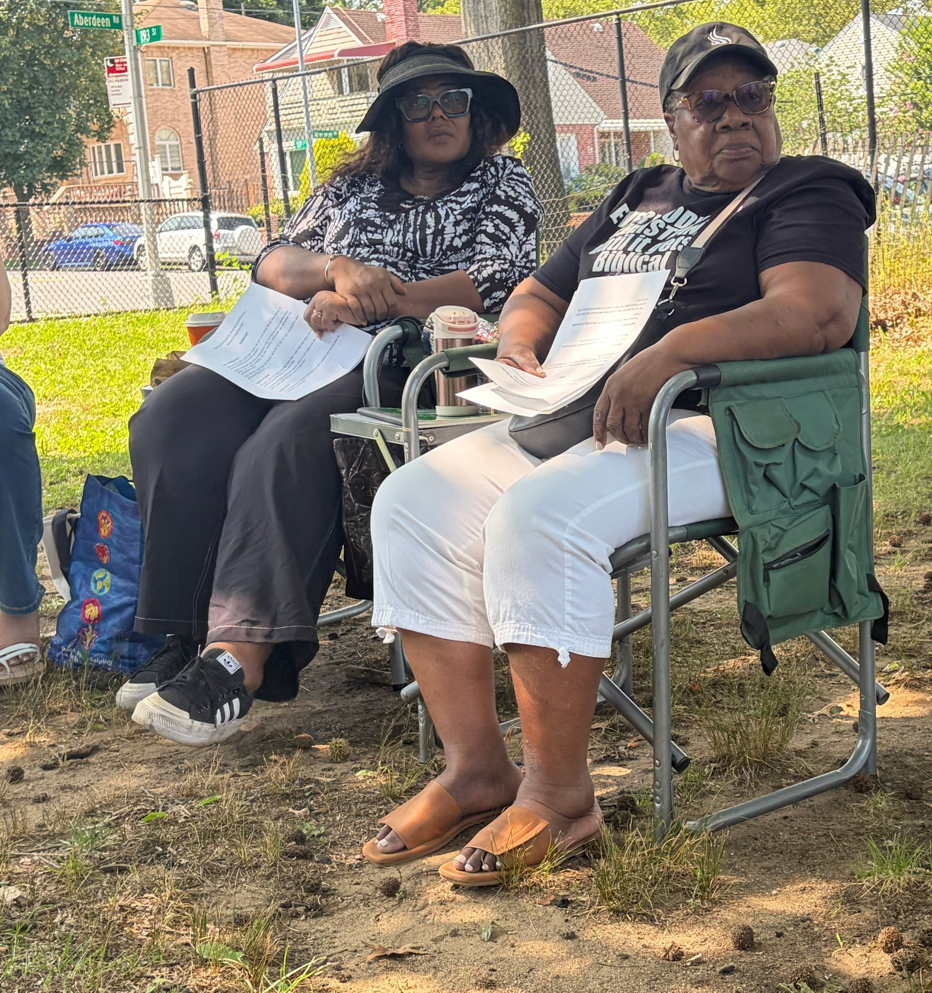 Two women seated outdoors on folding chairs in the shade, one holding papers, with a brick building and grass behind.