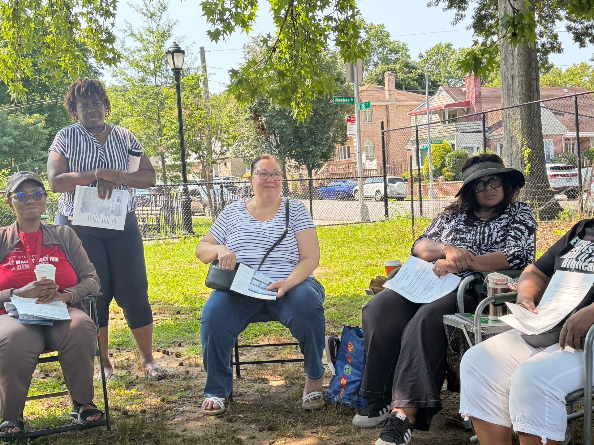 People seated outdoors in a park, holding papers, with trees, grass, and a playground in the background.