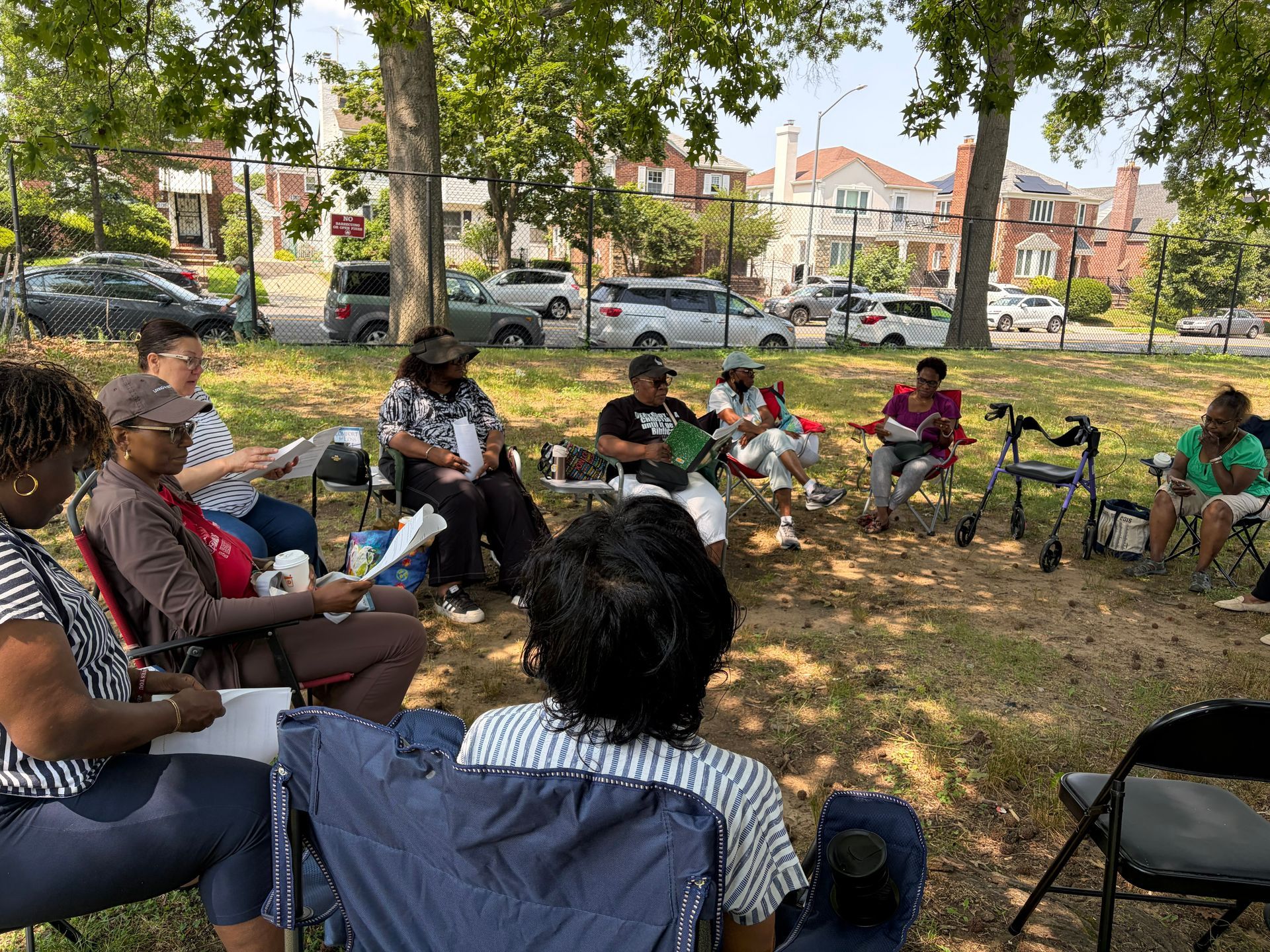 People sitting in a circle outdoors in a park, holding papers and speaking together under trees.