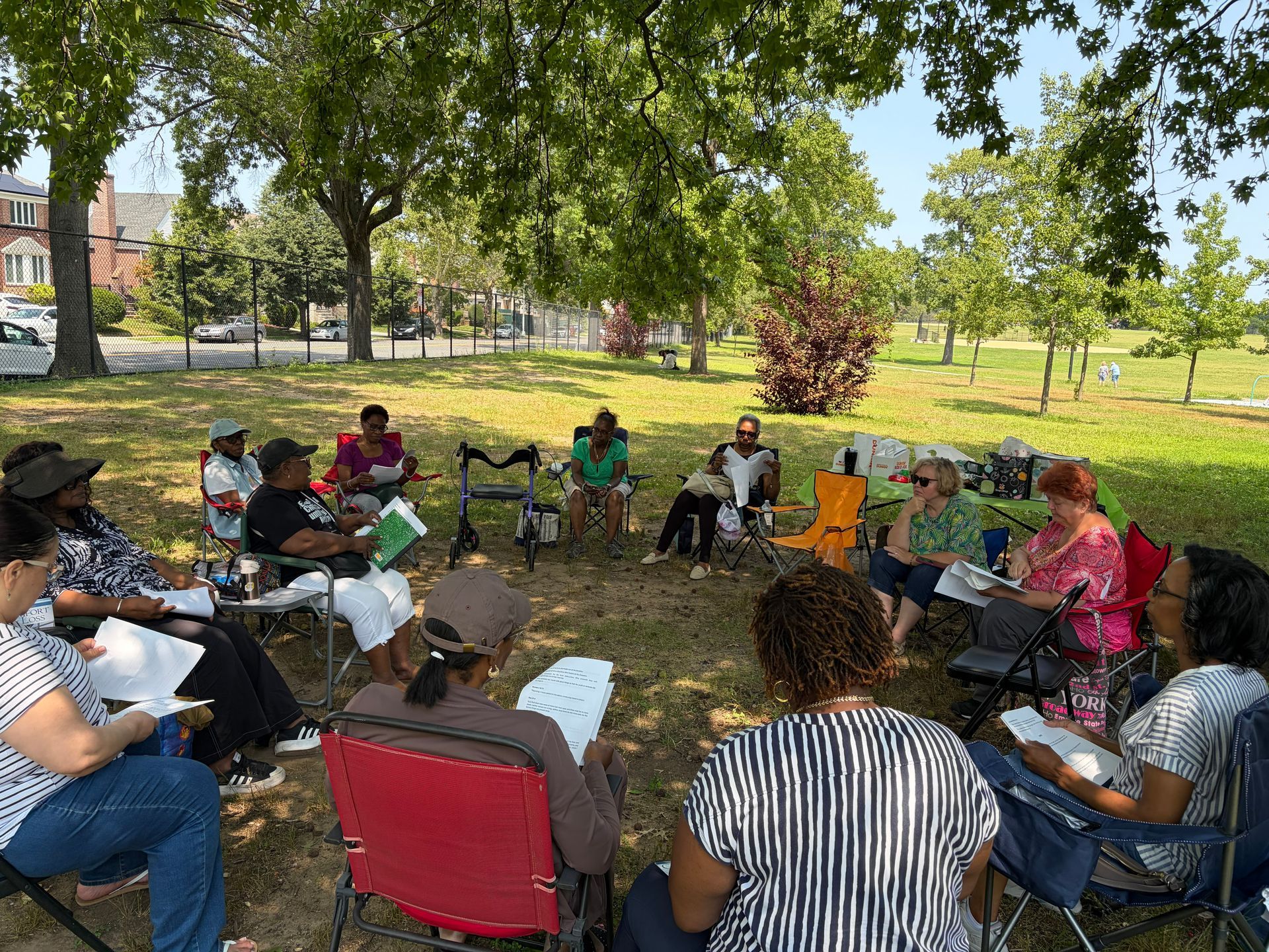 People seated in a circle outdoors under trees, holding papers and talking in a park.
