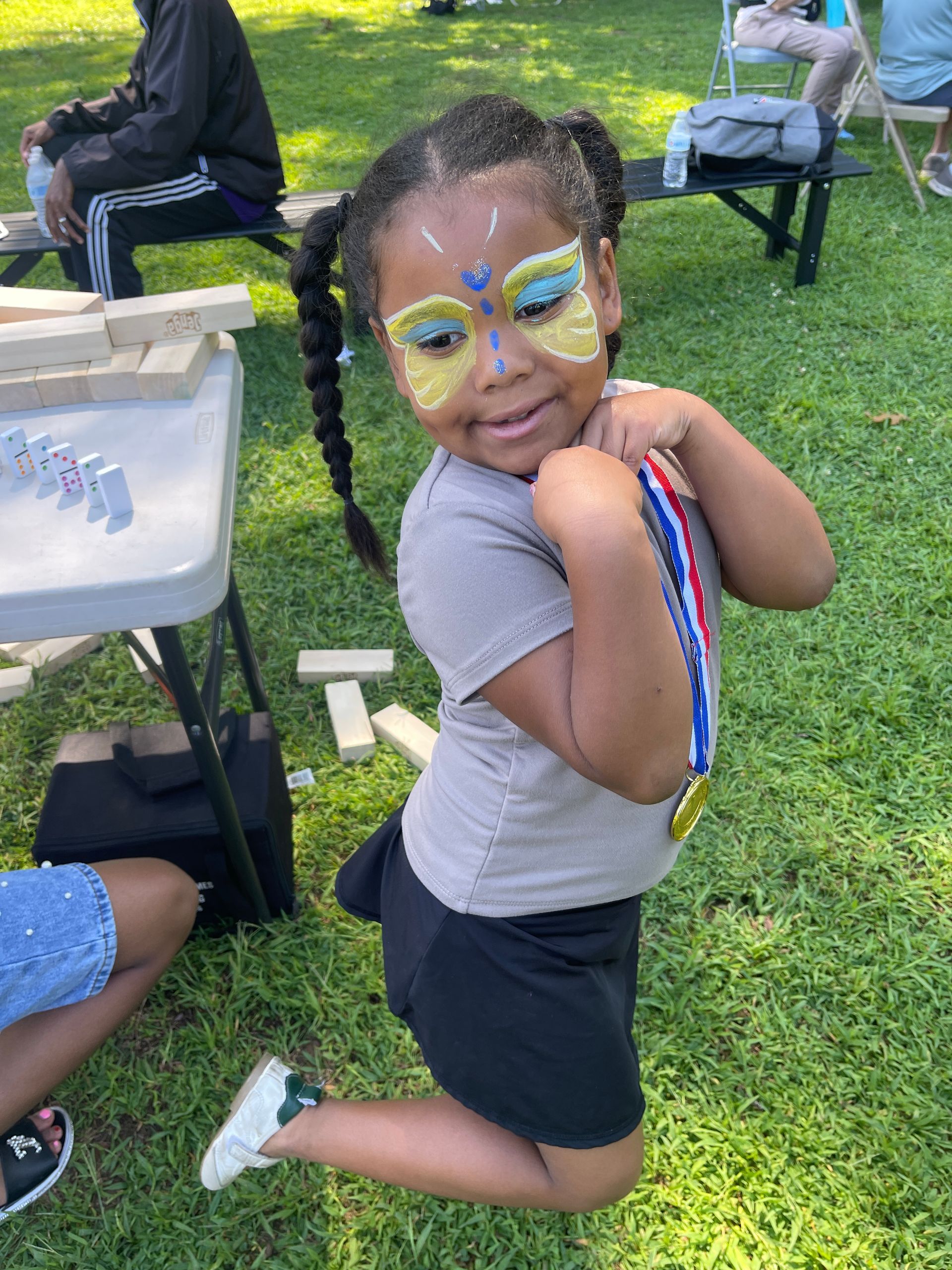 Child kneeling on grass with butterfly face paint and a beaded necklace at an outdoor event