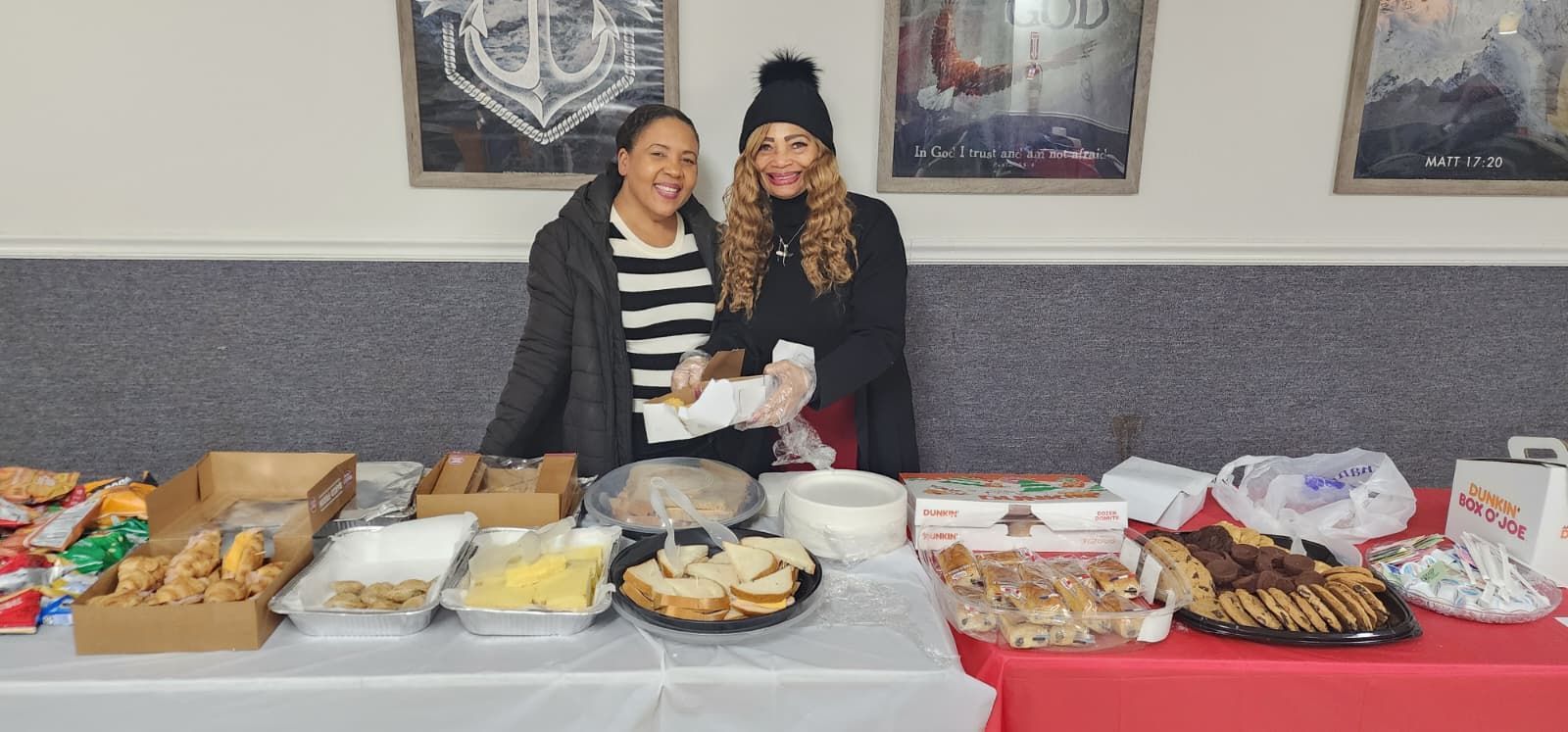 Two people standing behind a buffet table with trays of food and snacks in a room with framed wall art. Christmas Toy Drive 2025