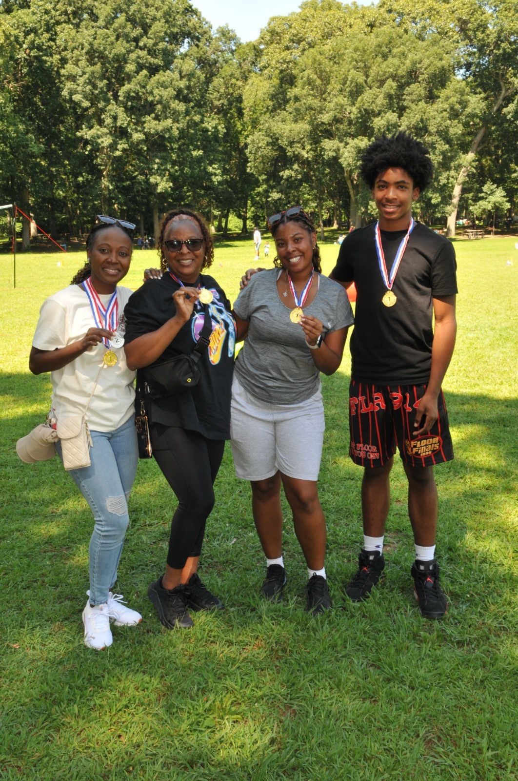 Four people standing on grass holding medals, posing outdoors in a park.