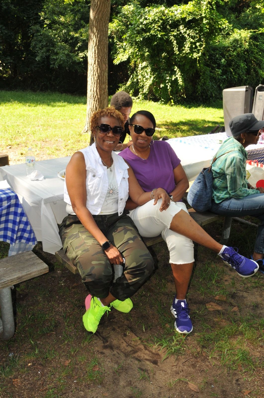 Two people sitting outdoors at a picnic table, smiling in sunglasses and colorful clothes.
