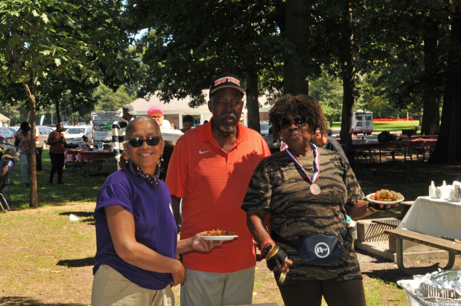 Three people posing outdoors at a picnic, with tables and trees in the background.