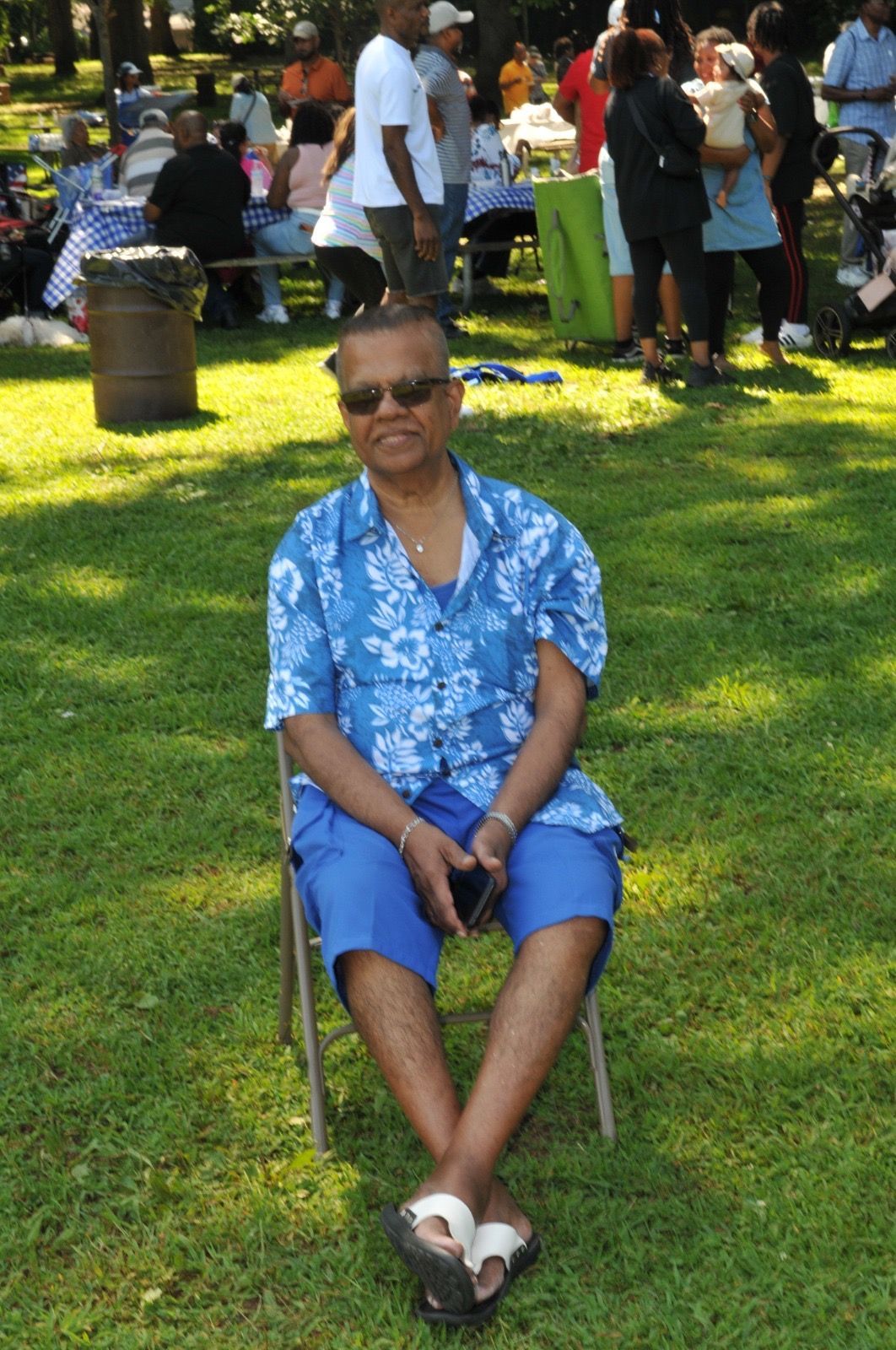 Person seated on a chair on a grassy lawn, wearing a blue floral shirt and sunglasses, with people gathered in the background.