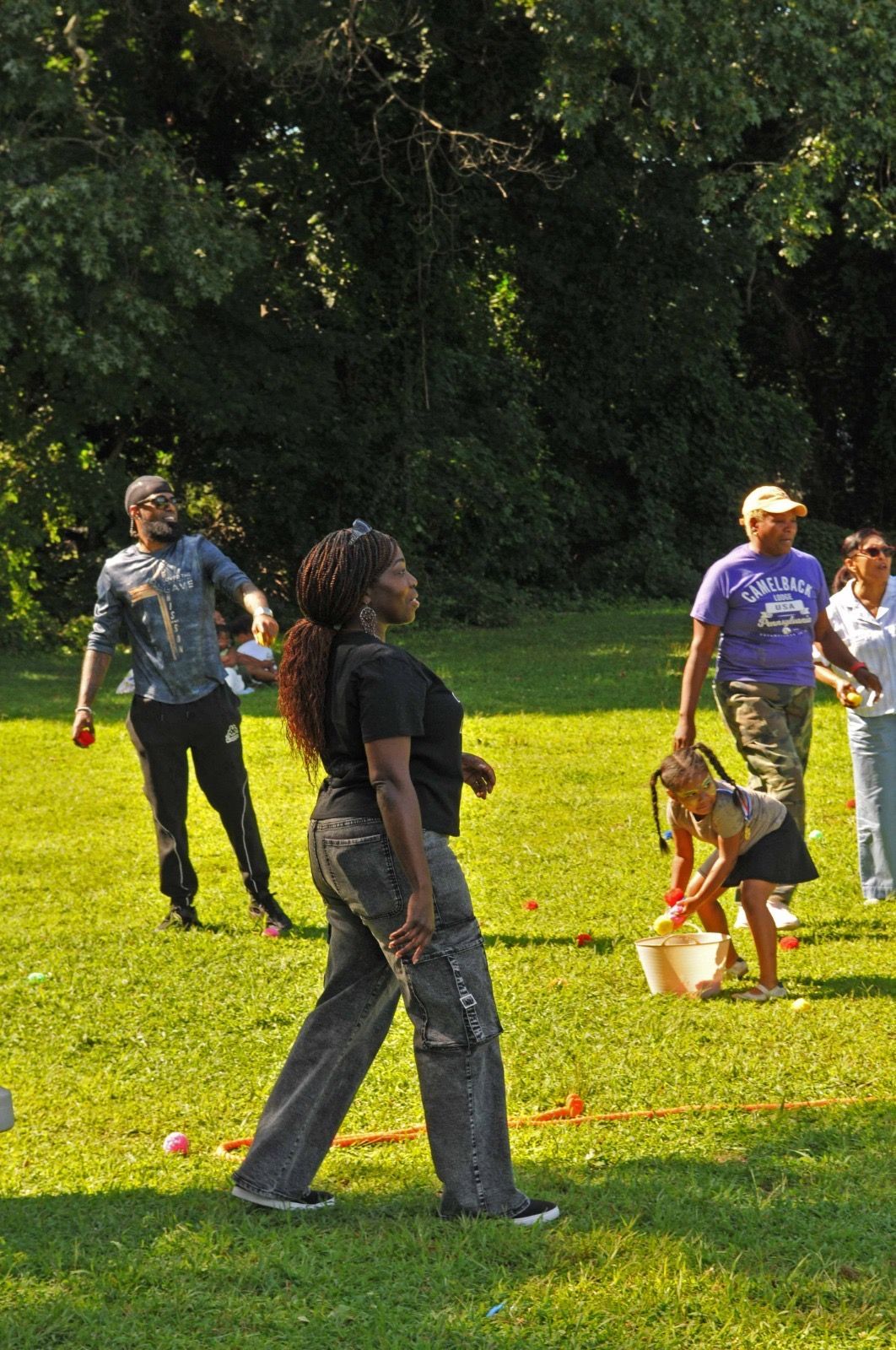 People standing and walking on a sunny grassy field near trees; one holds a small child’s hand.