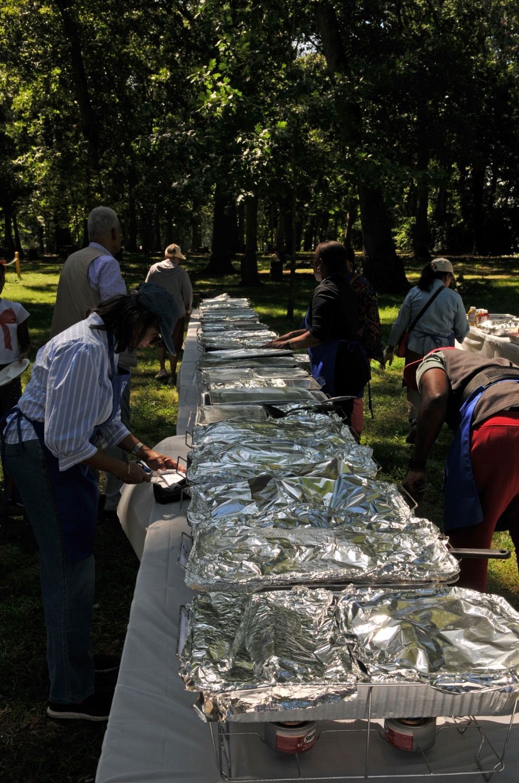 People serving food at a long outdoor buffet table covered with foil under trees