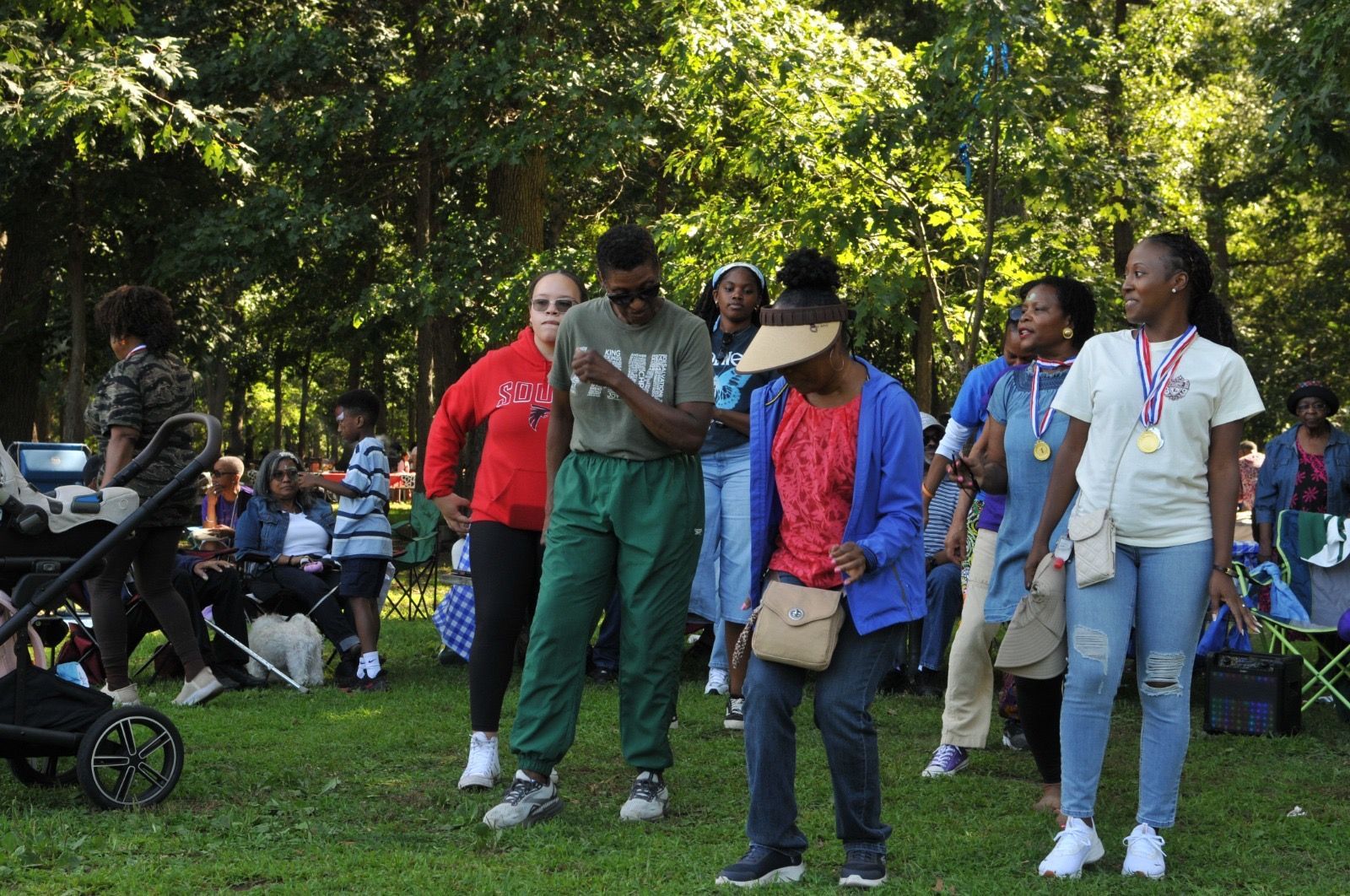 People gathered outdoors in a park, standing on grass near trees and strollers.